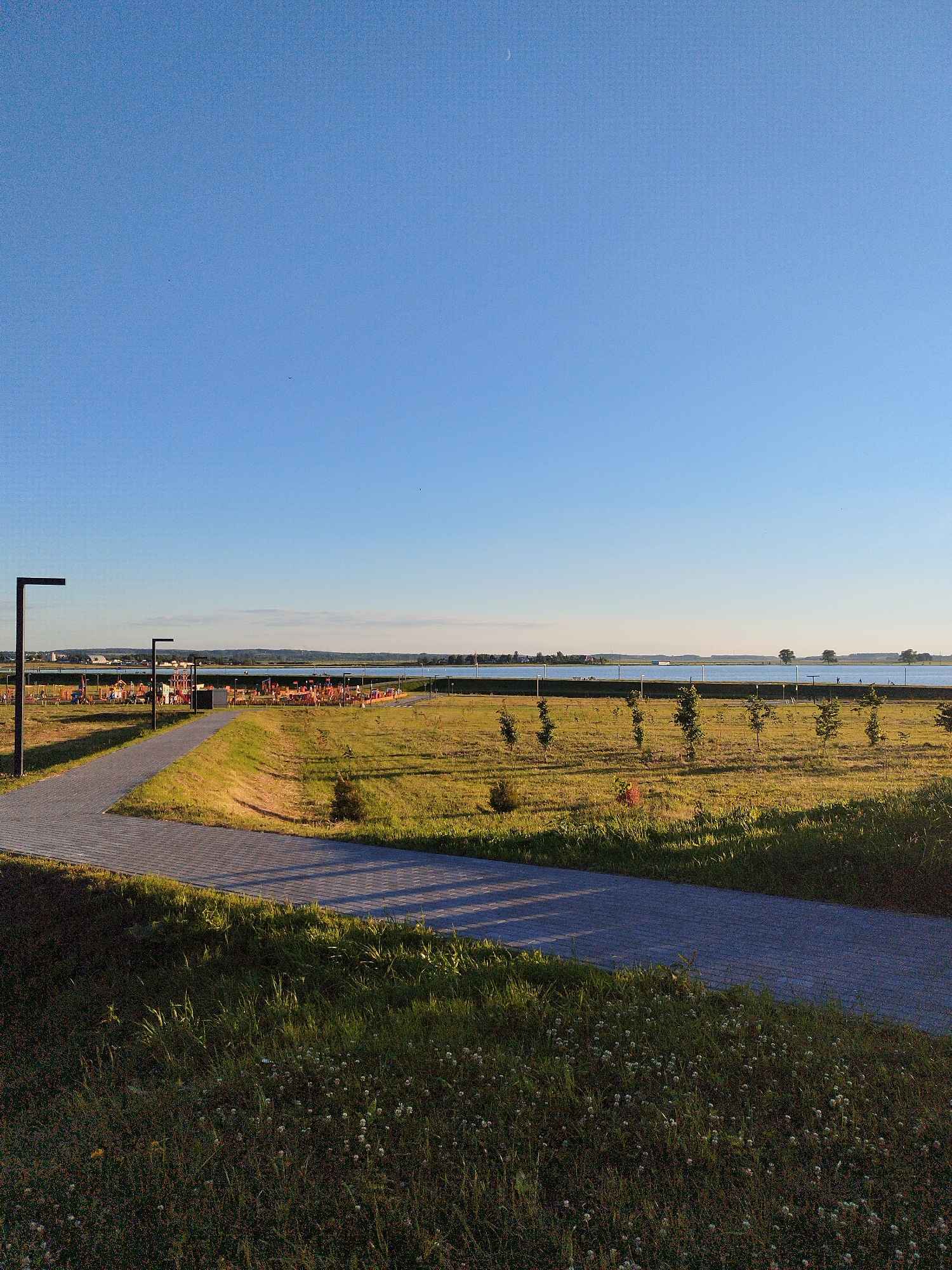 The image shows a public park with a paved walkway in the foreground, leading towards a body of water. The walkway curves from the lower left to the middle of the image. A tall, dark lamppost stands on the left side of the walkway. Beyond the walkway is a grassy field dotted with young trees. In the distance, across the water, there is a treeline and some buildings visible on the horizon. On the left side of the park area, there is a playground with colorful equipment. The sky is clear and blue, with a faint crescent moon visible in the upper right corner. The sun appears to be low in the sky, casting long shadows across the walkway and the grass. The overall scene depicts a serene and well-maintained outdoor recreational area.