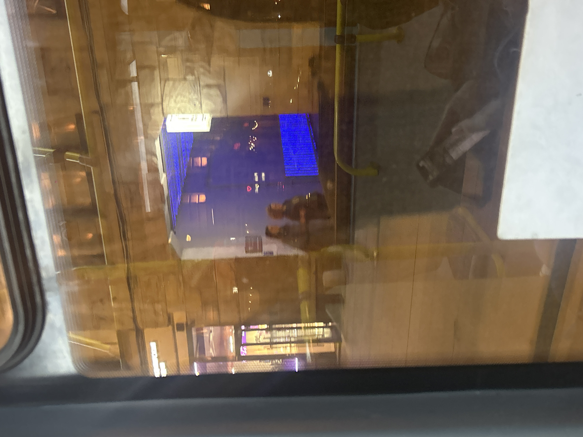 The media file presents a nighttime scene viewed through a window, likely from a vehicle such as a bus or tram, characterized by prominent reflections. The window glass has a warm, brownish tint, creating a layered visual effect.
The most striking feature is the reflection of a large building in the center, illuminated with numerous blue lights arranged in a grid pattern. This blue-lit structure dominates the view, suggesting a modern urban environment. Below this reflection, indistinct figures appear, also as reflections, with one person seemingly looking at a phone.
Reflected elements from the interior of the vehicle are clearly visible. These include a bright yellow horizontal grab bar and a yellow vertical pole on the right side of the frame. A white rectangular surface, possibly an interior panel or sign, is reflected in the upper right quadrant. The left side of the frame shows the dark edge of what could be a window frame or part of the vehicle's structure.
Looking through the reflections, the actual exterior environment appears dark and blurry, with hints of other distant lights or buildings in the background.
Visible text is present as a reflection on a horizontal sign or display in the lower middle portion of the image. Although somewhat blurry, it appears to be Cyrillic letters, most likely spelling "БАРСУК" (Barsuk), which means "Badger" in Russian. This could indicate a nearby business like a shop or cafe.
The image captures a dynamic urban nighttime scene in Saint Petersburg, Russia, with the interplay of interior reflections and external city lights creating a complex and atmospheric view.