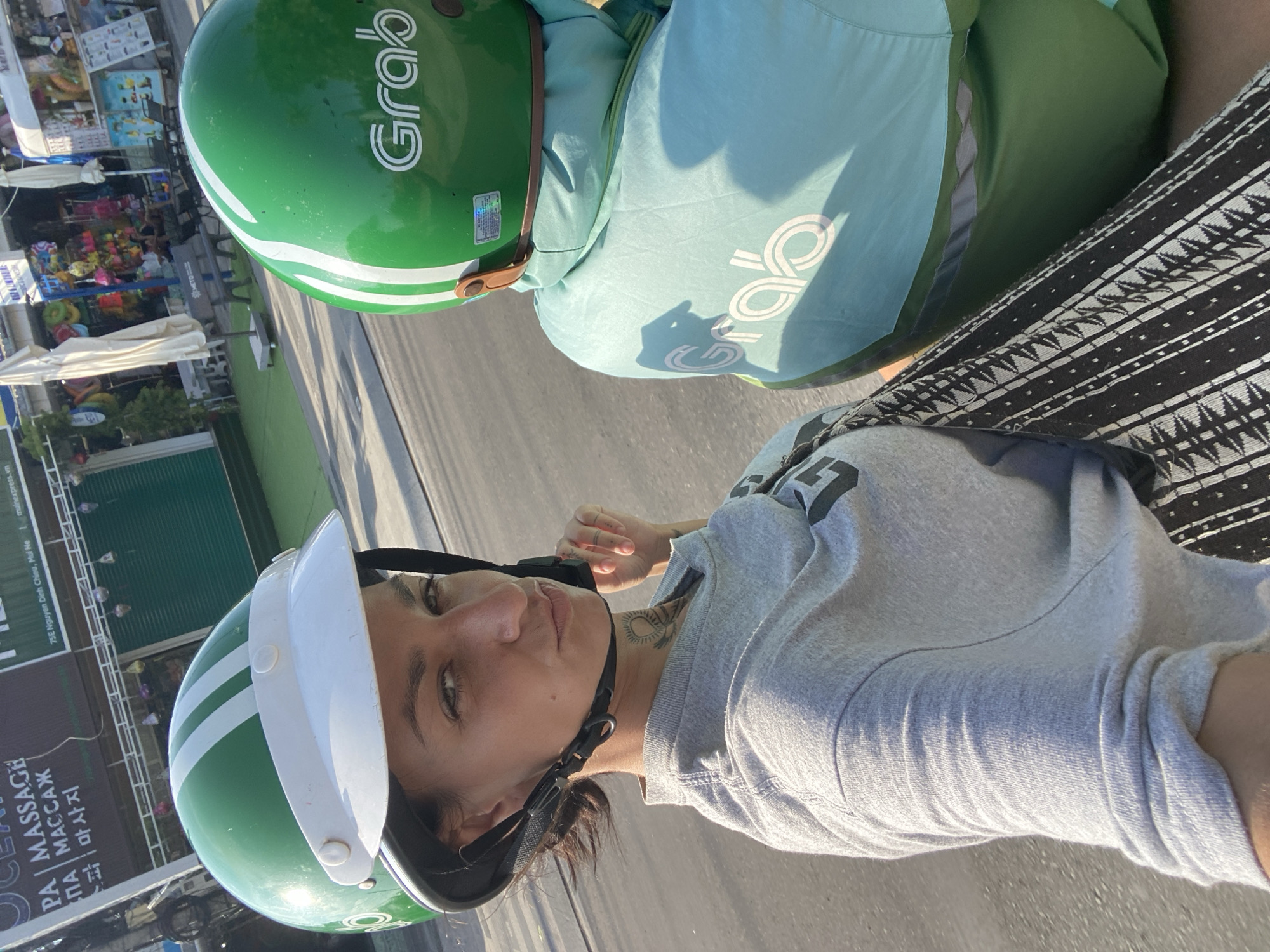 A woman in a grey t-shirt and patterned skirt rides on the back of a motorcycle, wearing a green helmet with white stripes. The motorcycle driver, also wearing a green helmet and a teal Grab jacket, is visible in the foreground. The background shows a street scene with shops, an awning, and the Grab logo visible on both helmets and the driver's jacket. The location is identified as Phu Thuy Ward, Vietnam. The lighting suggests it is daytime, with sunlight casting shadows.