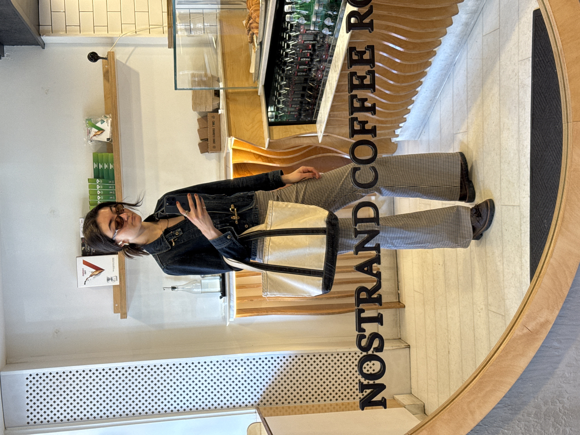 A mirror reflects a woman wearing sunglasses, a dark denim jacket, and patterned pants. She stands inside a shop called Nostrand Coffee Roasters, likely located in the city of New York, United States. The shop features shelves with various products, including coffee supplies and beverages. The woman is holding a bag. The mirror itself has the name of the store encircling its edges.