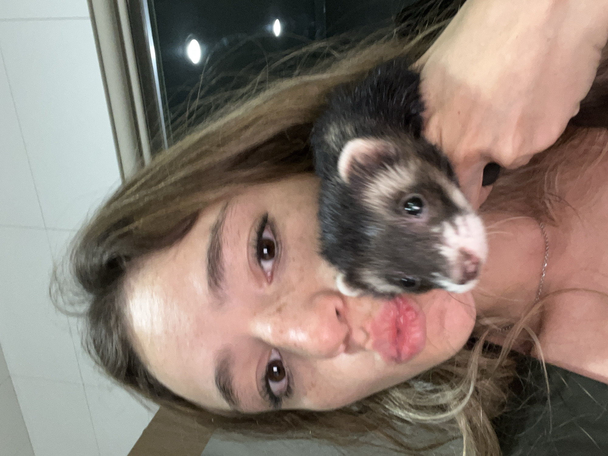 A selfie-style image captures a woman with long, light brown hair holding a ferret. The ferret's head is prominently featured, with dark fur and a white mask around its nose and eyes. The woman is making a kissing face. She has brown eyes, freckles, and a neutral skin tone. A silver necklace is visible on her neck. The background includes a window with an obscured view and overhead lighting. The location cannot be determined from this image.