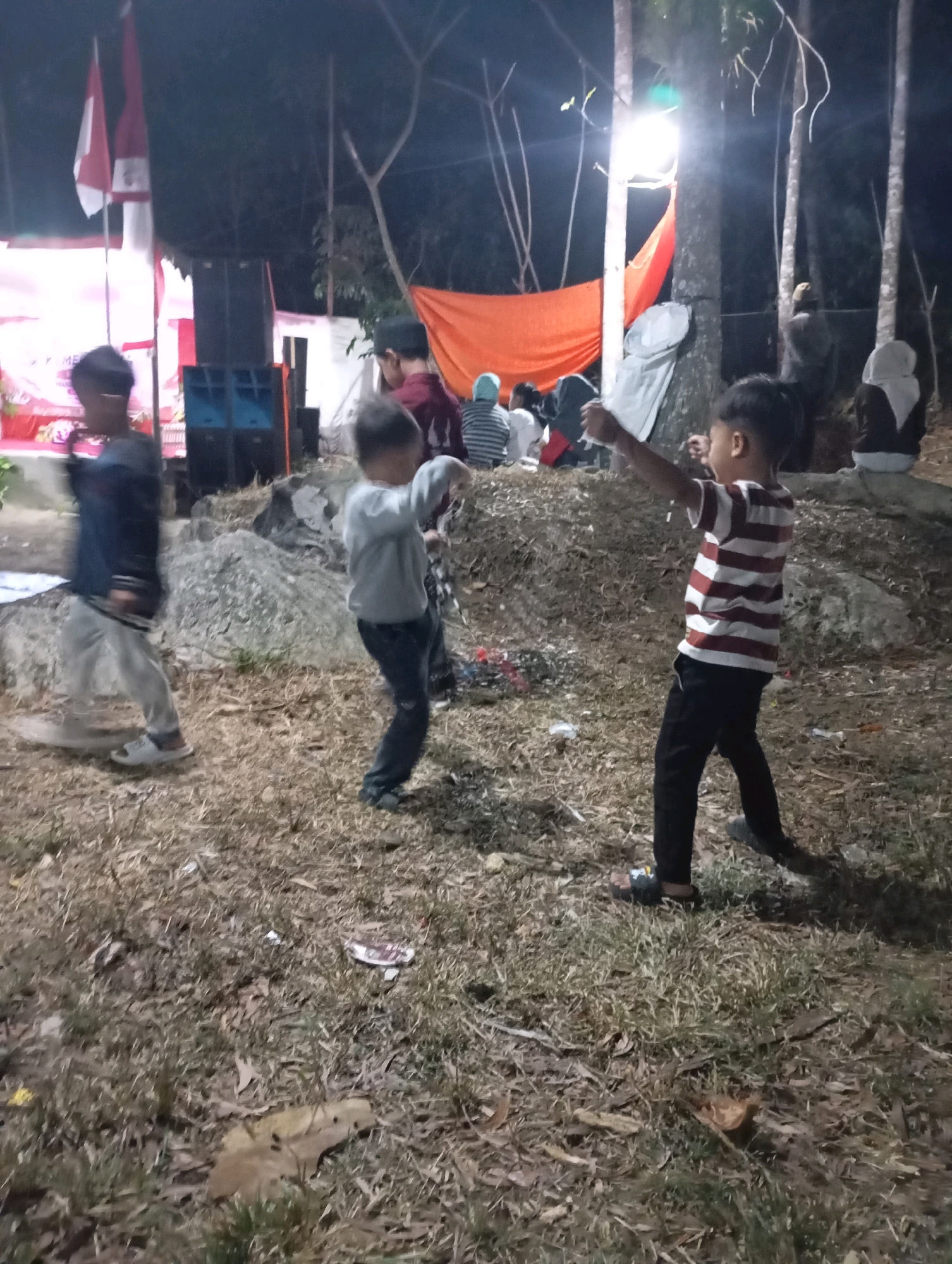 The image depicts an outdoor gathering at night in Ketepung, Indonesia. In the foreground, two young boys are visible. The boy on the right wears a horizontally striped red and white shirt and black pants, with his arms outstretched as if playing. The boy on the left wears a dark jacket and light-colored pants and is in motion, appearing to walk or run. Behind them, other individuals are gathered, some seated and others standing. A large orange tarp is draped over a structure, and stacks of speakers are positioned near a backdrop. Indonesian flags are also visible. The ground is covered with dry grass and scattered debris. The scene is illuminated by artificial lighting, casting shadows.