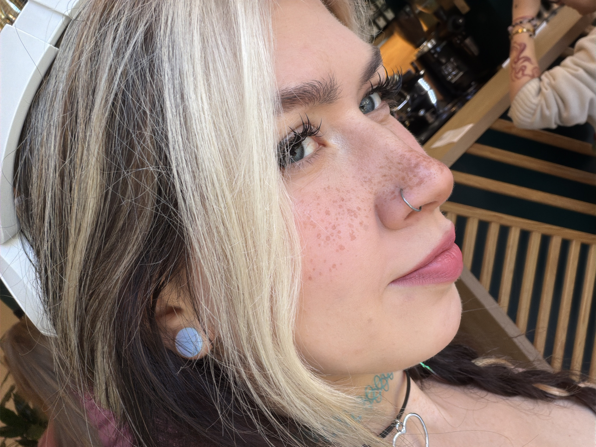 The image depicts a close-up, slightly angled portrait of a young woman with heterochromatic eyes, one appearing bluish-gray. She has fair skin with prominent freckles across her nose and cheek. The woman has a silver nose ring in her left nostril. Her hair is a two-toned style with dark brown at the roots transitioning into blonde streaks. She has visible false eyelashes. A blue, disc-shaped earring is visible in her left ear. The woman wears a thin black choker necklace with a silver heart-shaped pendant. There is a teal-colored tattoo visible on the side of her neck.
The background is partially blurred, revealing what appears to be a café or restaurant setting with dark wooden furniture and metal objects that suggest kitchen equipment. A person wearing a light-colored sweater is partially visible in the background, with a decorated arm suggesting body art. The image could have been taken in an outdoor setting, with wooden beams and a slatted wall visible. There is no concrete evidence to place the location specifically in Budva, Montenegro.