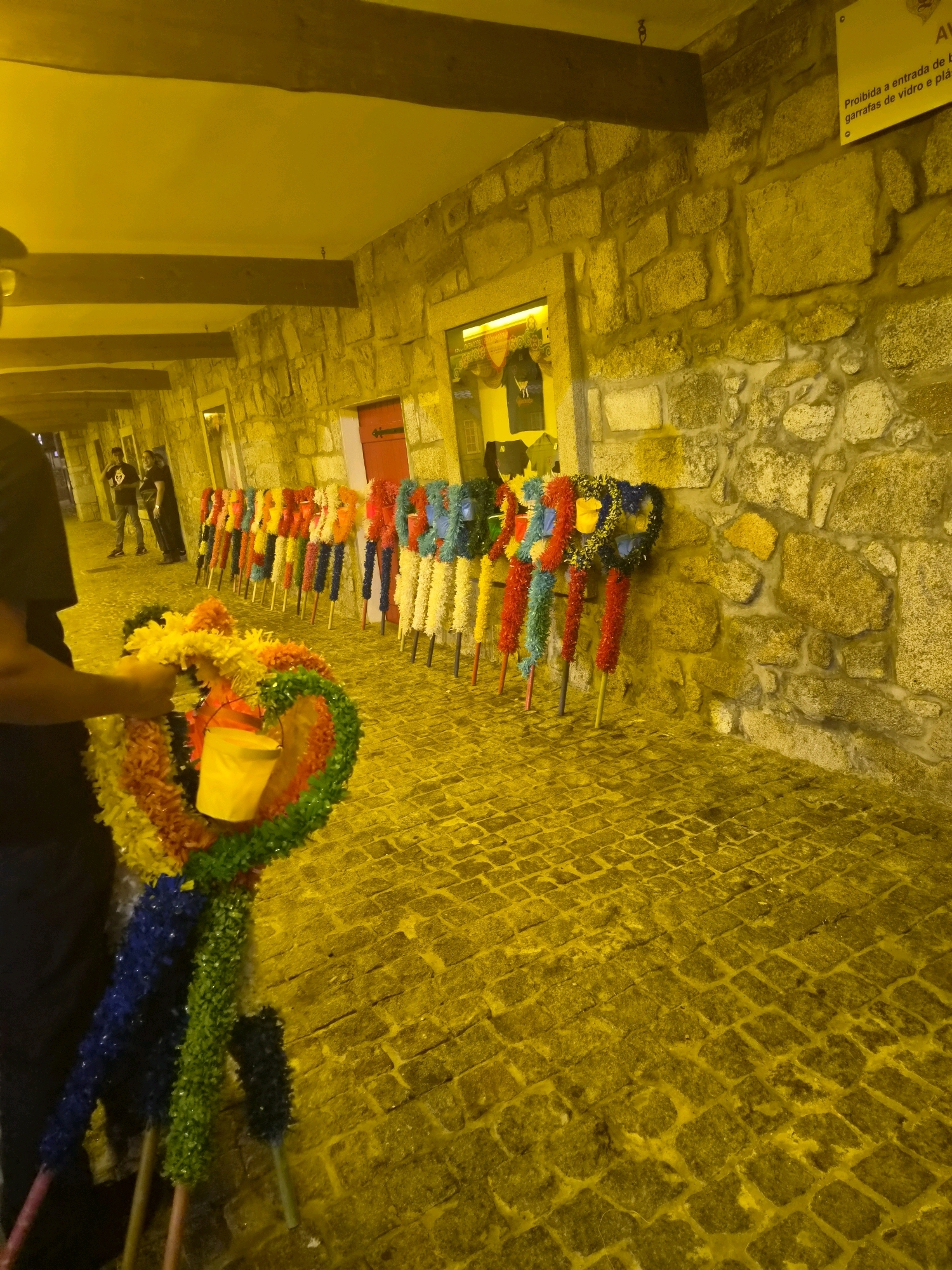 A stone-walled arcade with a cobblestone floor in Darque, Portugal, is filled with colorful, festive decorations. In the foreground, a person in dark clothing is holding a large, heart-shaped wreath made of vibrant tinsel in shades of red, orange, yellow, green, and blue. Behind them, a row of similar decorations, each mounted on a wooden pole and adorned with colorful tinsel, stands against the stone wall. These decorations vary in design, with some featuring a more floral or fan-like appearance. To the right, a sign with Portuguese text, including "Proibida a entrada de bebes...," is affixed to the wall. The overall scene suggests a celebratory or festive event, possibly a local festival or market.