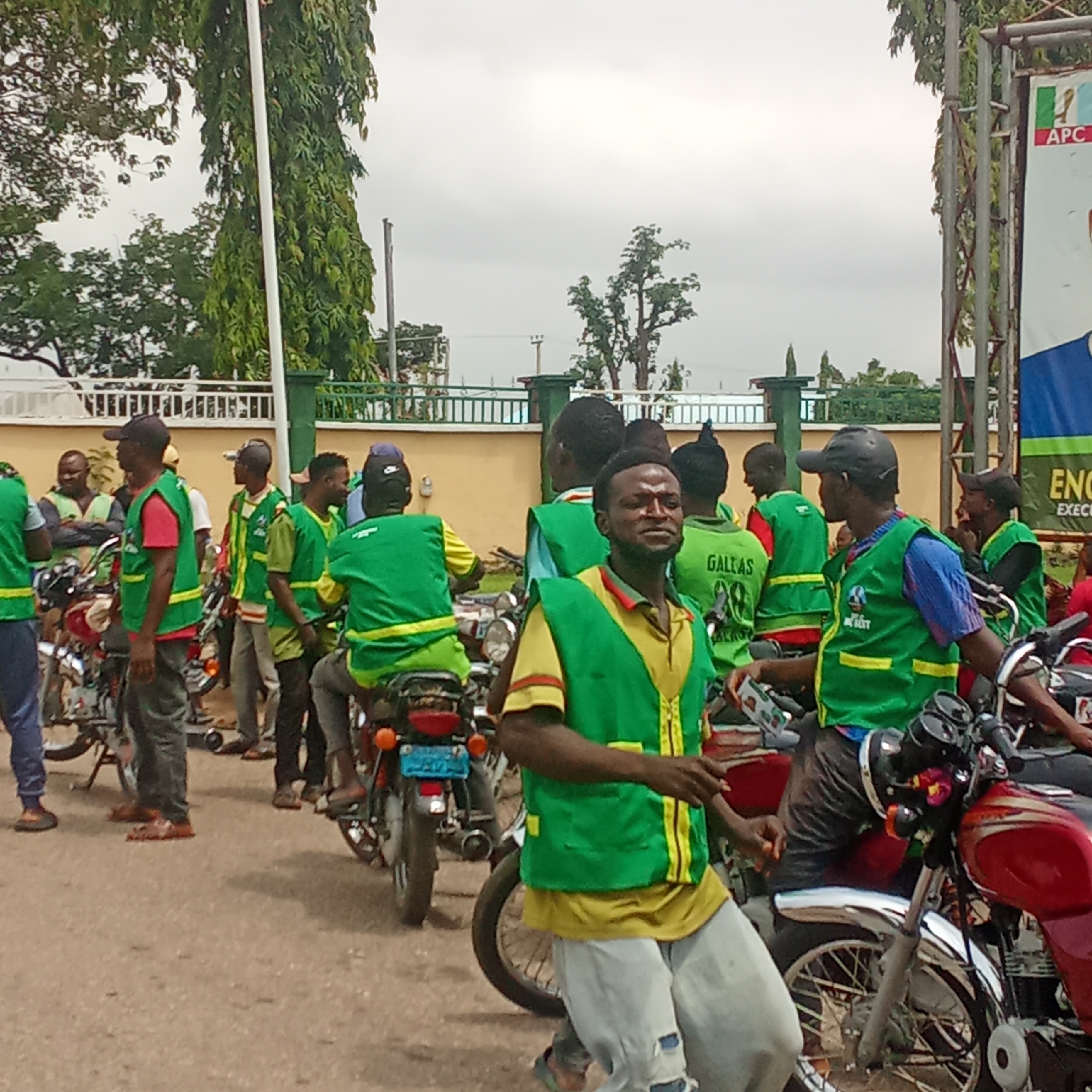 The image shows a group of men wearing green and yellow vests standing beside motorcycles on a street. A building with a green-topped fence is in the background. A sign with the letters "APC" is visible. The men are congregated on the street. The location is identified as Akwanga, Nigeria.