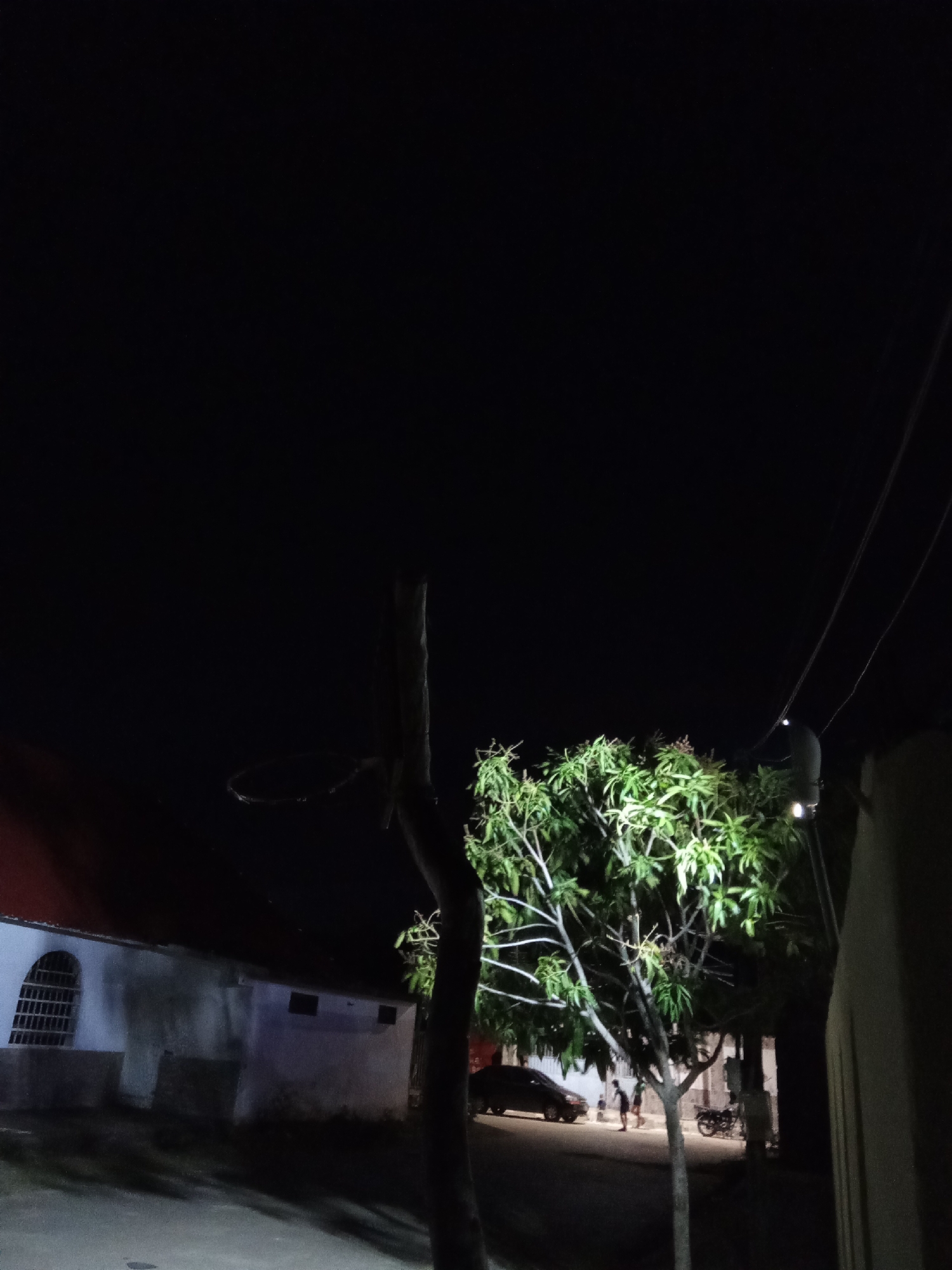 This is an outdoor nighttime scene in Juan Griego, Venezuela. The image shows a street illuminated by an overhead light source, possibly a streetlight, which highlights a tree with green leaves.
To the left, a building with a red roof and a barred window is visible. A basketball hoop without a net is mounted on a pole in front of the building.
Further down the street, there's a parked car and two people walking. A motorcycle is also visible. Overhead power lines are strung across the sky.
The foreground features a tree trunk, and the overall scene is dark, with details visible due to the artificial light.