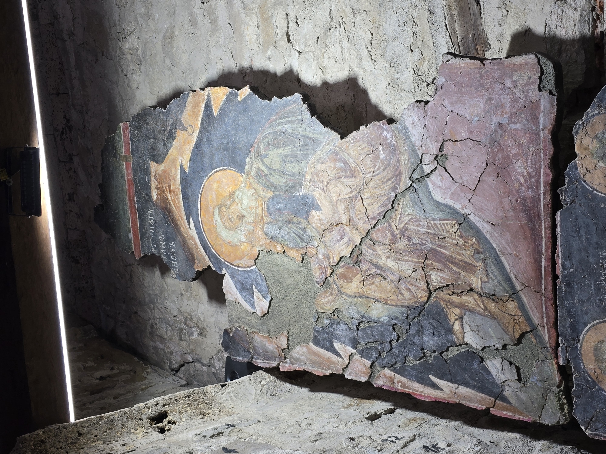 The image shows deteriorated sections of wall frescoes in an indoor setting. The frescoes appear damaged and fragmented with visible cracks and missing portions. The setting seems to be a room or structure made of stone, with rough, uneven walls. A light source is present, casting a stark glow on one side of the scene. The image does not contain enough information to identify the exact location or the city. People are not visible, and the subjects of the frescoes are religious figures.
