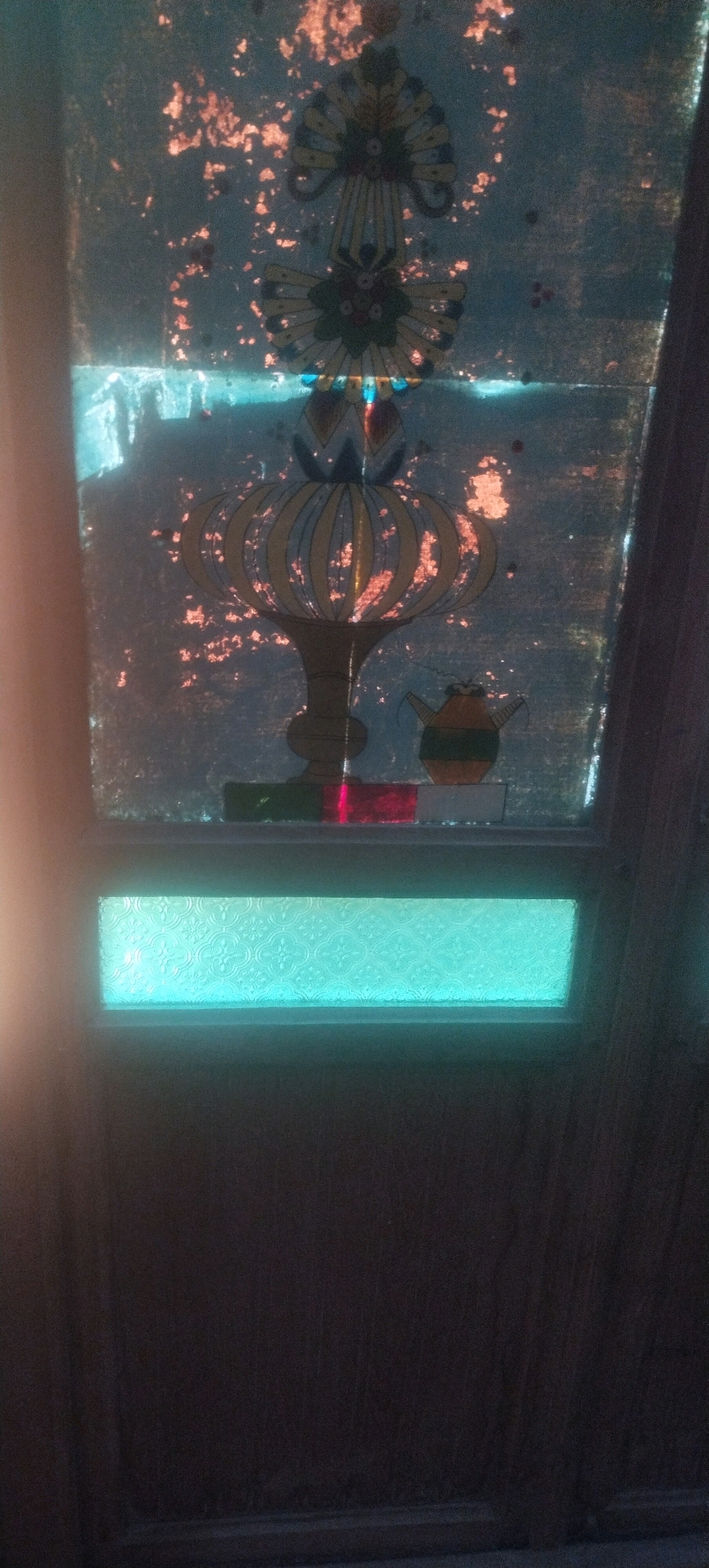 The image displays a stained glass window with an ornate design featuring floral and vase-like motifs. The glass is a mix of blue, yellow, and red, with scattered orange-red particles that may be rust or damage. Below the stained glass, there is a horizontal pane of textured, light blue glass. The rest of the image shows the dark brown, wood-grain texture of a door frame. There are no people or animals visible in the image, nor any indications of a specific location in Daleman, Indonesia. The scene appears to be an indoor setting, possibly a room with a door and a window.