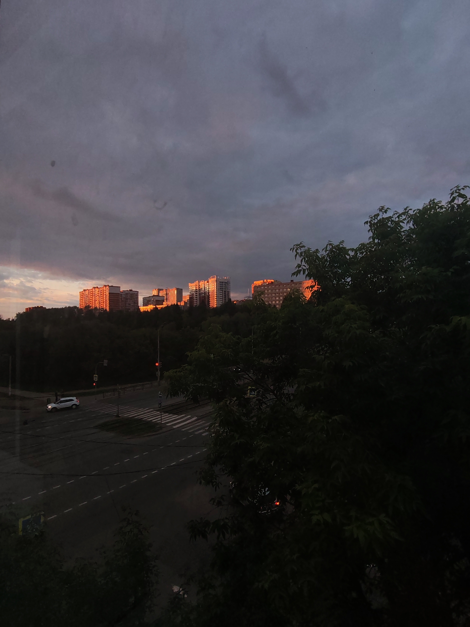 The image depicts a twilight scene in Izhevsk, Russia, overlooking a residential area. In the background, several multi-story apartment buildings are illuminated by the setting sun, casting a warm orange glow on their facades. The buildings are situated on a hill covered with dense dark green trees. In the foreground, a street intersection is visible with a pedestrian crossing. A white car is turning at the intersection, and a traffic light is visible with a red signal. The sky is overcast with dark gray clouds, with hints of pink and orange from the sunset. The overall atmosphere is calm and somewhat somber due to the fading light and dark foliage.