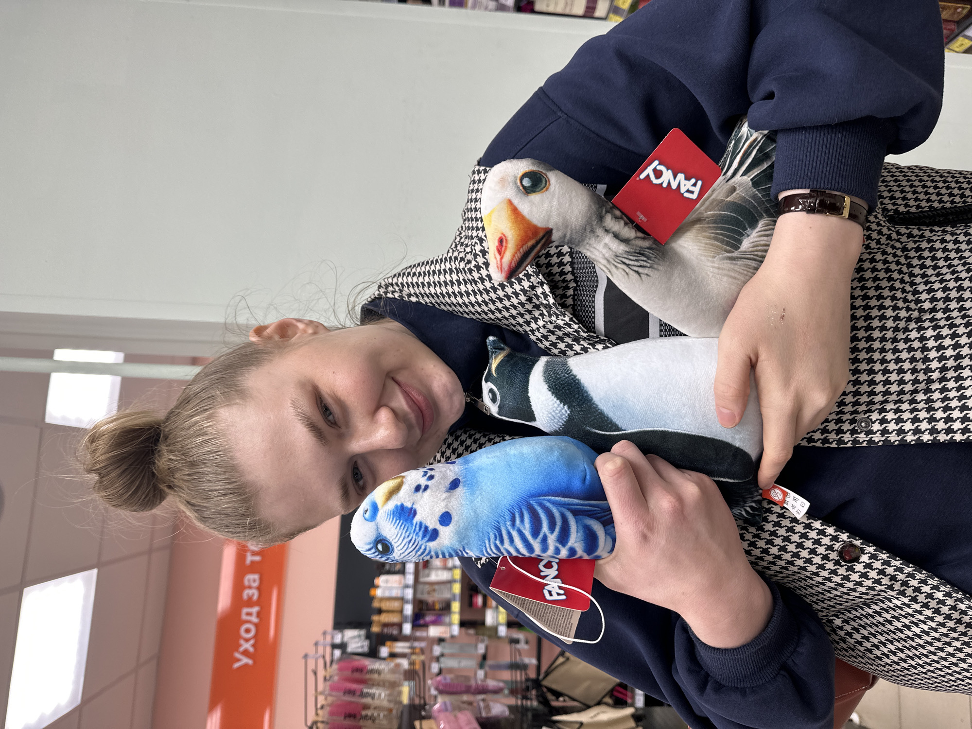 A young woman is holding three stuffed birds while standing in a store. The birds are a goose, a penguin, and a parakeet, each with a "FANCY" brand tag attached. The woman is wearing a navy blue sweater and a black and white houndstooth patterned jacket. She has her hair pulled back in a bun. The store appears to be a retail environment with shelves stocked with various items visible in the background. The sign "Уход за т" (Care for T) in Russian is visible on an orange panel. Based on the store signs in Russian, this image likely originated in the city of Kazan, Russia.