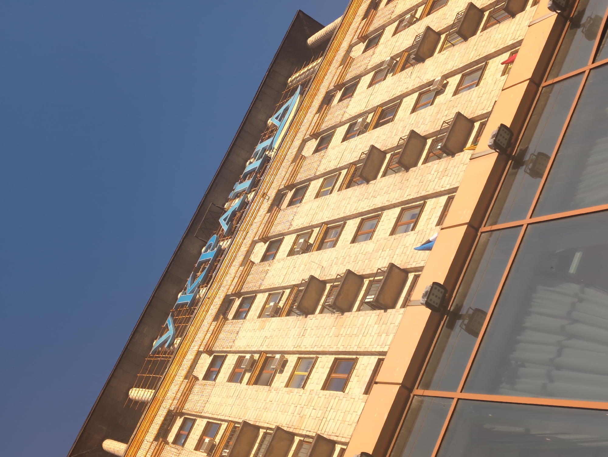 This is a low-angle, outdoor photograph of the Ukraina hotel in Kyiv, Ukraine, taken on a clear, sunny day. The building, constructed of light-colored bricks, dominates the frame, with a deep blue sky visible above. The hotel's name, "УКРАЇНА," is spelled out in large, blue, three-dimensional letters across the top of the facade. The architectural style is evident with numerous windows, balconies, and air conditioning units visible on the side of the building. At the bottom of the image, a section of a modern glass-fronted structure with orange framing is visible, reflecting the sky and some interior details. There are no people visible in the photograph.