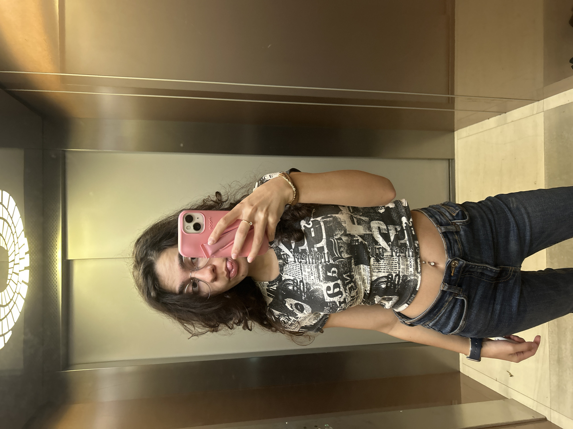 A young woman is taking a selfie in an elevator with a silver and beige interior. She is wearing jeans, a cropped black and white patterned top, eyeglasses, a bracelet, and a smartwatch. She is holding a pink phone with an "eponti" branded case. The elevator has a circular light fixture visible in the mirror. Given the description, it is not possible to pinpoint the exact location of the elevator in Sofia, Bulgaria, without more specific information.