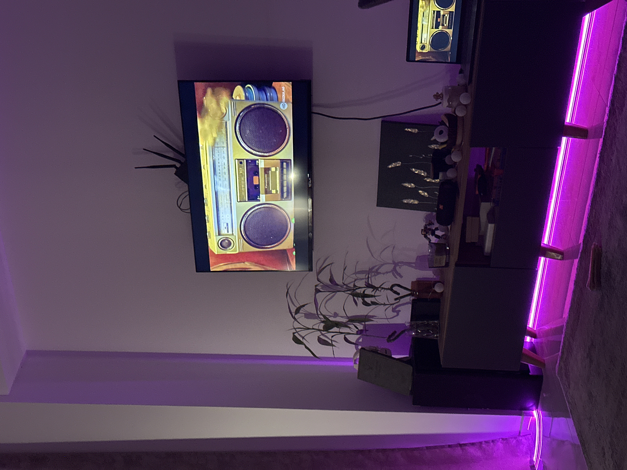 The image depicts an interior space with a television mounted on the wall, displaying an image of a boombox. Below the TV, there's a dark-colored TV stand with decorative items and a plant on it. The room is illuminated by purple LED lighting, which runs along the baseboard and highlights the side of the TV stand. In the upper part of the image, another TV is seen, displaying an image of a boombox. The location is Budva, Montenegro, a city known for its coastal setting and historic architecture.