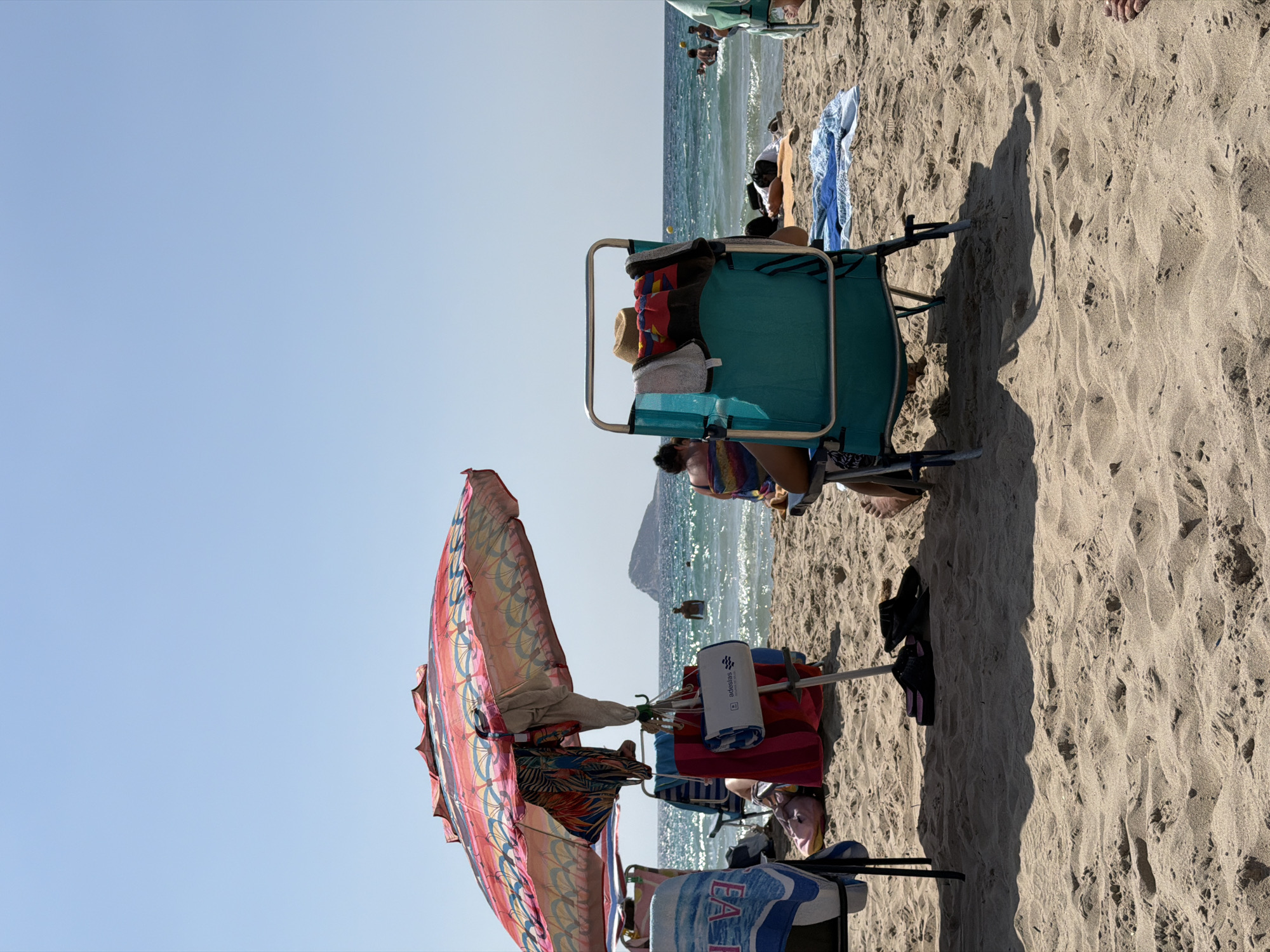 The image depicts a sunny day at a beach in Urbanització Cales I Talaies, Spain. The scene is set on sandy ground with the blue ocean and a distant coastline visible in the background. Several beach chairs are set up, some occupied by people. In the foreground, a brightly colored beach umbrella with a pink, yellow, and blue geometric pattern is open, casting a shadow on the sand. A green beach chair is also visible, with towels and a straw hat draped over it. Another person appears to be sitting in a chair near the water. The sky is clear and blue, indicating pleasant weather. The presence of beach umbrellas, chairs, and people in swimwear suggests a recreational activity, likely a day of relaxation and enjoyment at the beach. There is text visible on a white cylindrical object on a chair which reads "adaslas". The overall mood appears to be relaxed and summery.