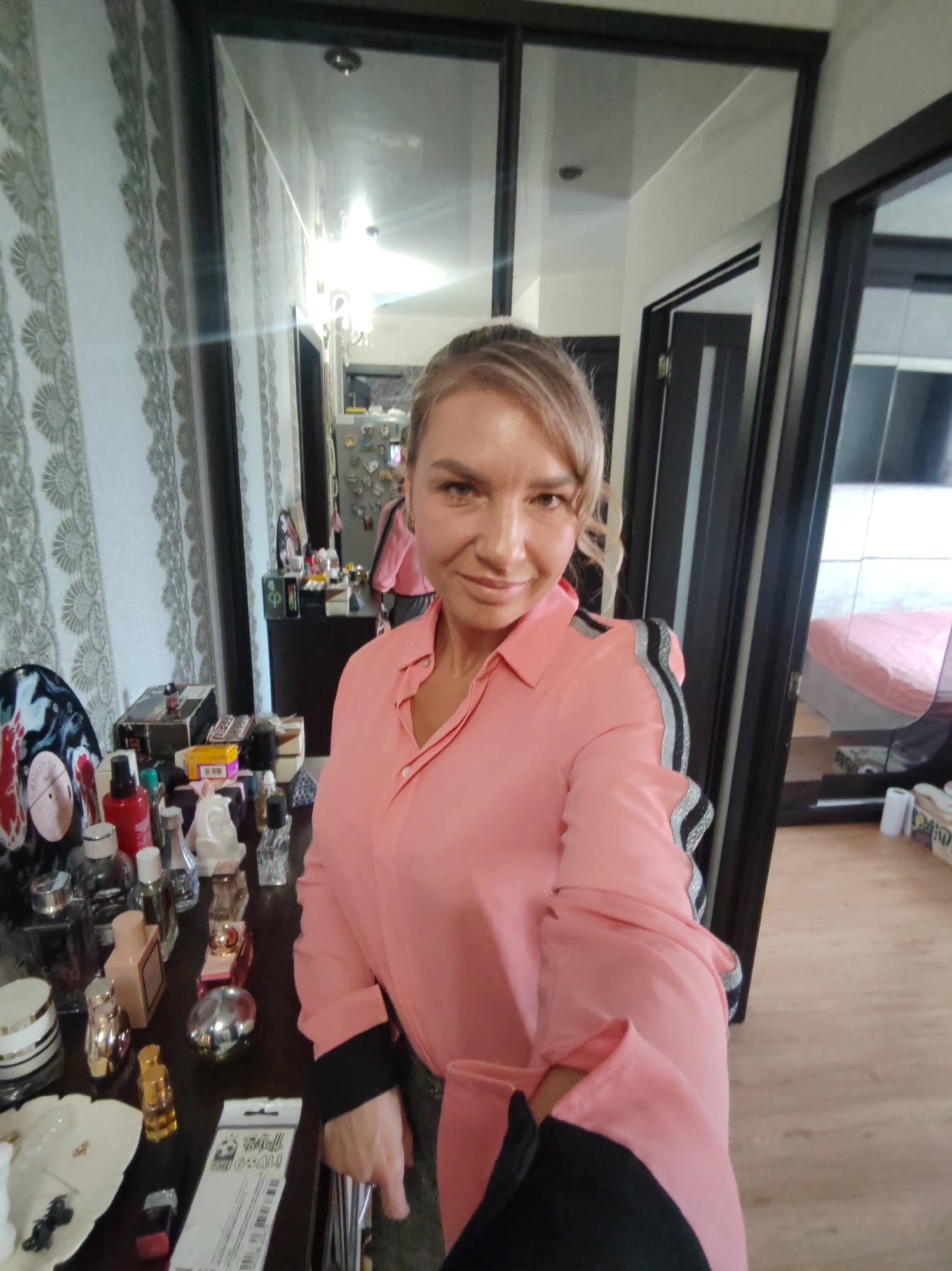 A blonde woman in a pink collared shirt stands in a room, taking a selfie. She is positioned in front of a table filled with various perfume bottles and cosmetics, with a large mirror reflecting the room behind her. To her left, a patterned wallpaper is visible, and to her right, a closet door and a bed with pink bedding are reflected in the mirror. The overall setting appears to be a domestic interior, possibly a bedroom or dressing area, in Petropavlovsk-Kamchatsky, Russia.