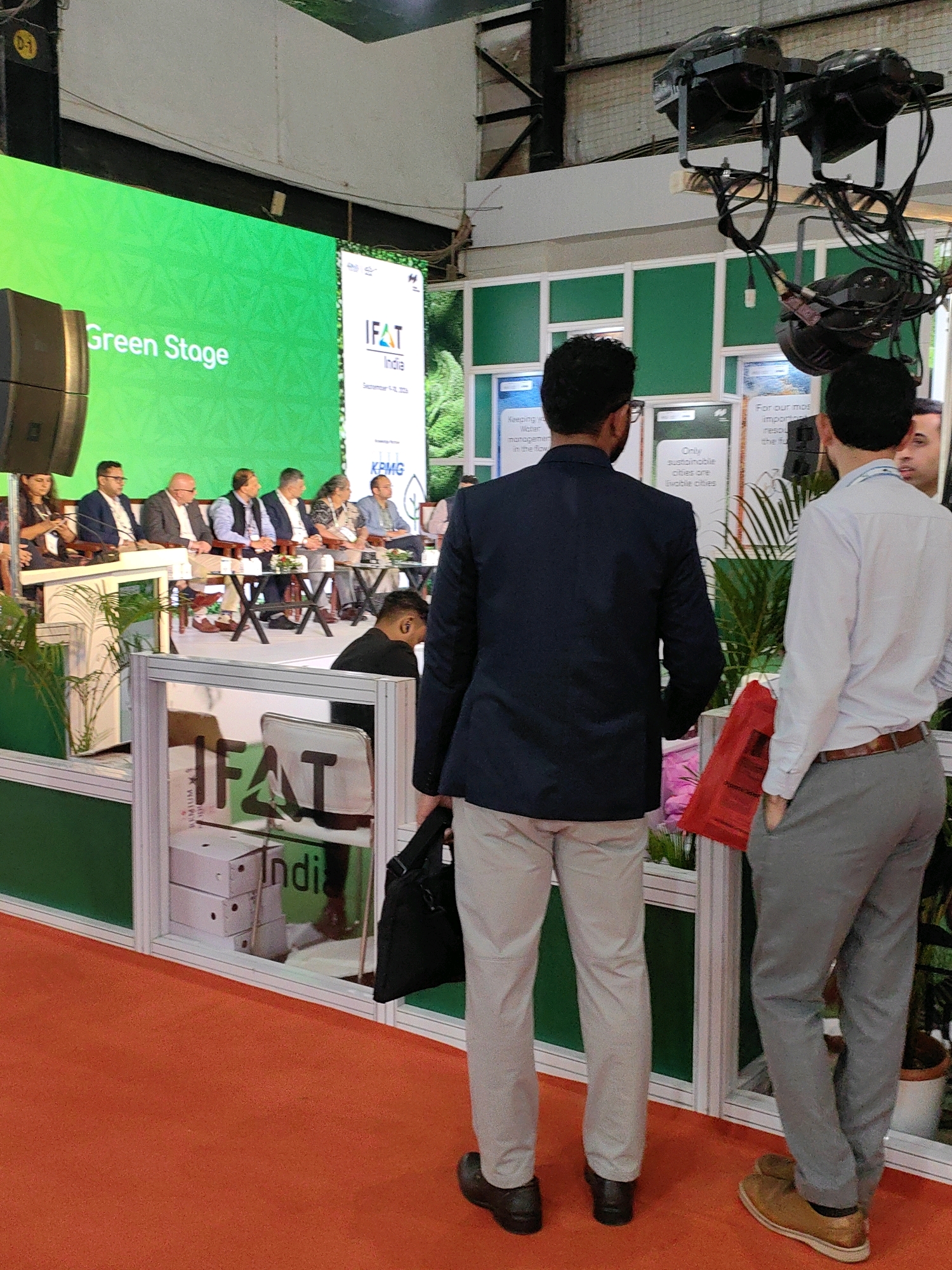 In Mumbai, India, at an exhibition hall, a panel of seven individuals is seated on a raised stage in front of a large green screen displaying "Green Stage." To the right of the stage, two men stand facing away from the camera, engaged in conversation. The man on the left wears a navy blue suit jacket and light grey pants, while the man on the right wears a light blue collared shirt and grey pants. Between them, a bright red bag is visible. In the foreground, a glass-enclosed display features boxes and what appears to be the logo "IFAT India." The floor is covered in an orange carpet. Overhead, industrial lighting fixtures are suspended from the ceiling. The environment suggests a professional event or conference.