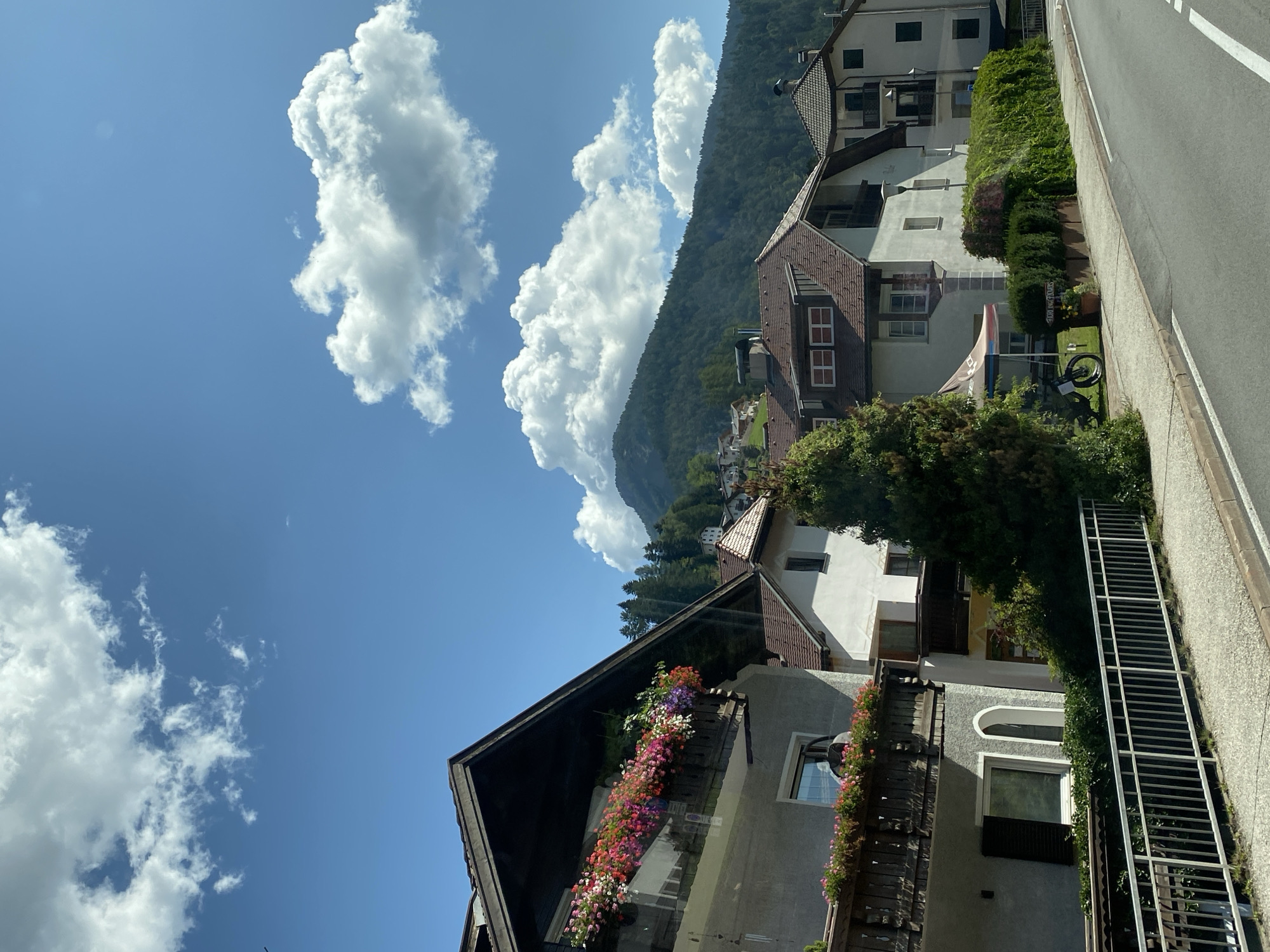 The image shows a street scene in the city of Urtijëi - St. Ulrich in Gröden - Ortisei, Italy. In the foreground, a paved road runs diagonally from the bottom right to the middle left of the frame. A light-colored sidewalk runs parallel to the road, separated by a white line. A white metal railing borders the sidewalk.
On the left side of the street, a multi-story building with a gray facade and a dark brown roof dominates the foreground. It features balconies adorned with abundant pink and red flowers. Below the main balcony, another balcony with fewer flowers and a railing is visible. A window with dark shutters is also present on this level.
Adjacent to this building, other residential structures with light-colored facades and brown roofs are visible. Some have dark balconies, and several have windows of various sizes. One building features a red-tiled roof and appears to have multiple levels. A tall, dark green evergreen tree is positioned in front of these buildings.
A blue and white tent with the word "BIKE" visible is set up on the sidewalk next to a dark-colored electric bicycle. A small potted plant sits near the tent.
In the background, a lush, green, tree-covered mountainside rises steeply. The mountain is partially obscured by white, puffy clouds against a bright blue sky. A few scattered houses are visible on the lower slopes of the mountain. The overall impression is a sunny day in a picturesque mountain town. No people are visible in the image.