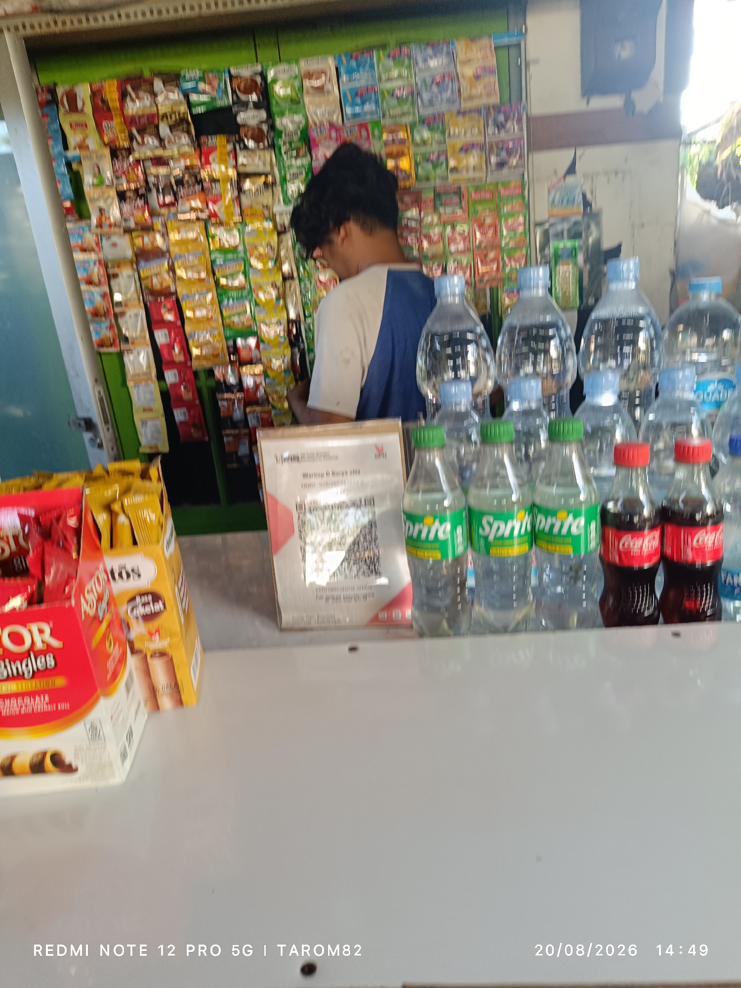 A person with dark, curly hair is standing in front of a shelf filled with various snacks and drinks. The person is wearing a white shirt with blue accents on the sleeves. The shelf is stocked with numerous small packets of food products, mostly in colorful packaging, and several bottles of beverages. In the foreground, there are several rows of plastic water bottles, some with green caps and others with red caps. Among these are also bottles of Sprite and Coca-Cola. To the left, a box of "Astor Singles" chocolate wafer rolls is visible. A QR code scanner is placed on the counter. The setting appears to be a small shop or convenience store. The timestamp in the lower right corner indicates "20/08/2026 14:49".
