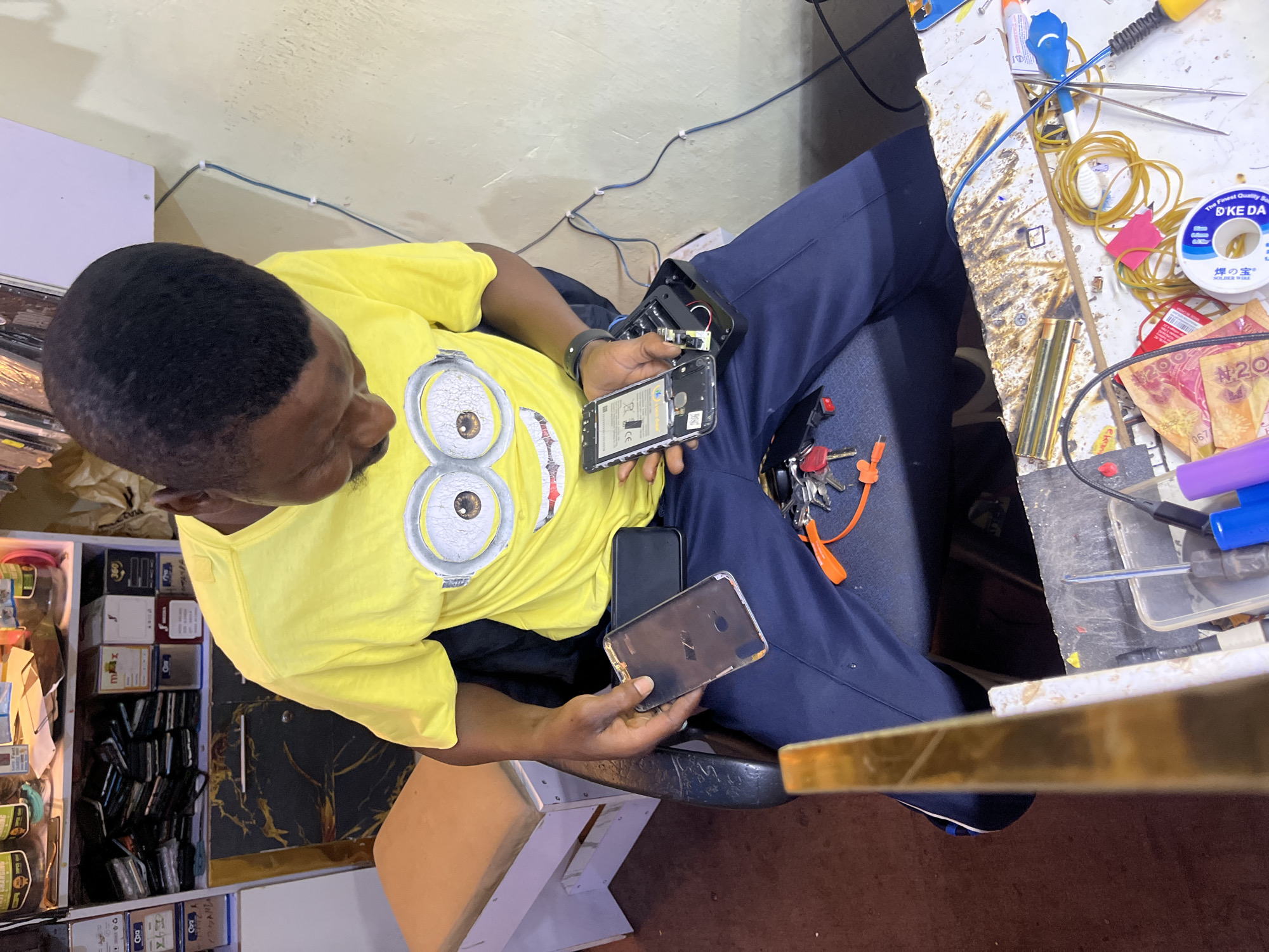A man wearing a yellow t-shirt with a Minion graphic sits on a chair in what appears to be a workshop or repair shop. He is holding a disassembled smartphone in his hands, with some of its components scattered on the desk in front of him. The desk is cluttered with various electronic repair tools and supplies, including wires, rubber bands, a soldering iron, and what looks like a roll of solder wire. There are also several mobile phones visible in a shelf unit to the left of the man, along with boxes and jars. The man is focused on the phone he is holding, suggesting he is in the process of repairing it. The lighting suggests it is daytime, and the overall setting indicates a busy workspace. Visible text on the solder wire spool includes "DKE DA" and Japanese characters, and on some money bills, "#20" and "067" are visible.