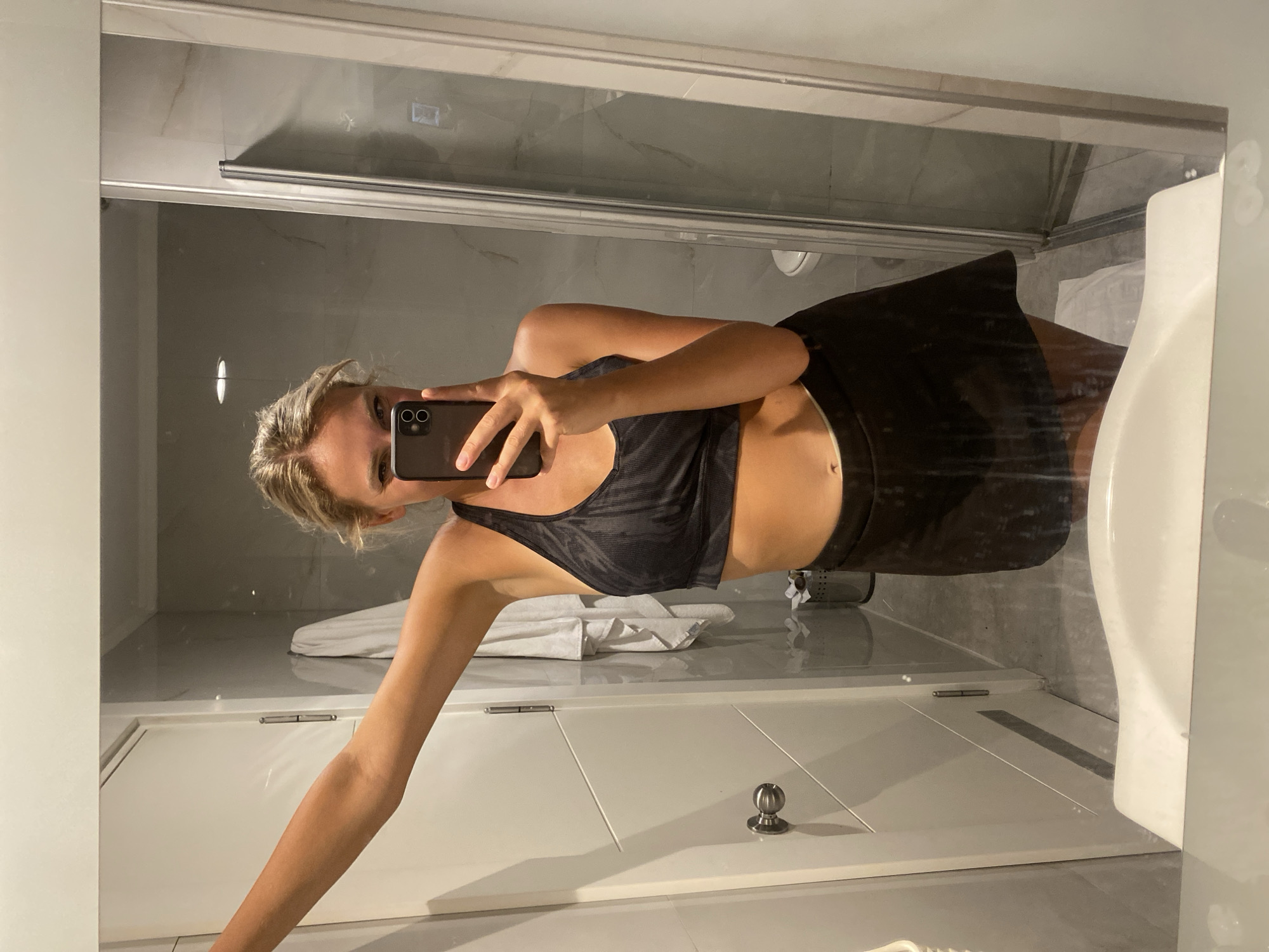 The image is a selfie taken in a bathroom, likely in Aksu, Turkey, based on the request. A young woman with blonde hair pulled back is holding up a smartphone to take the picture. She is wearing a black sports bra and a black skirt. Her left arm is extended, leaning against the mirror. The bathroom features a white sink in the foreground, a white door with a silver handle on the left, and a shower enclosure with a glass door on the right. The walls appear to be tiled with a marble-like pattern. A white towel hangs on a rack near the door, and a trash bin is visible near the shower. The lighting suggests it is indoors, possibly in a hotel room.