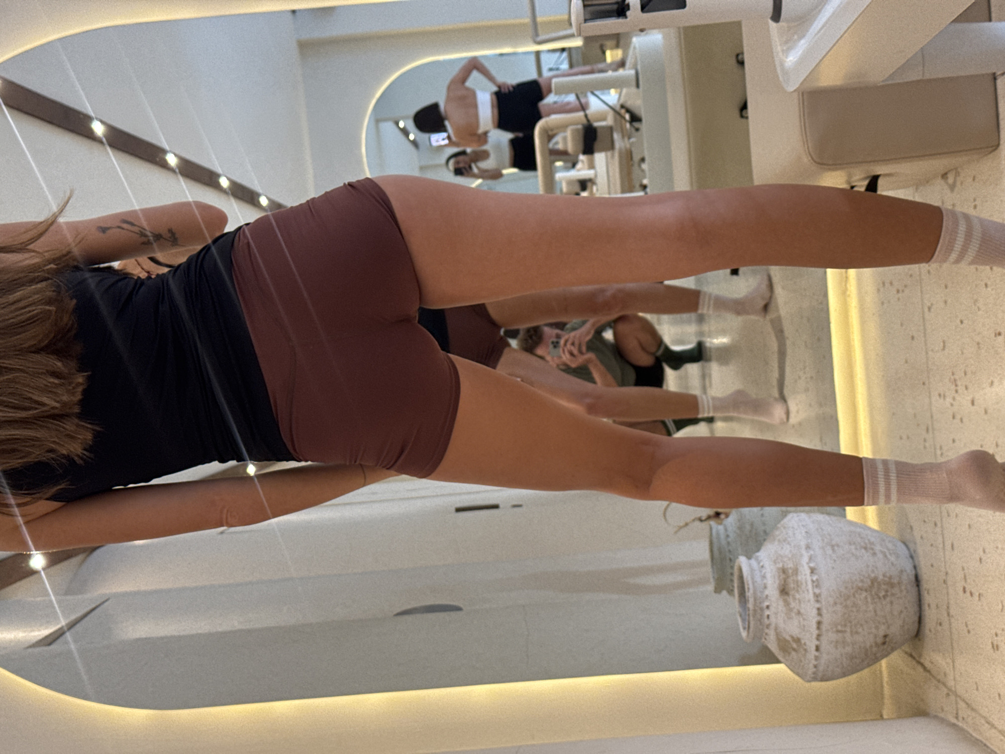 The image is a reflection in a mirror, showing a woman in athletic attire, consisting of a black top and brown shorts, wearing white socks. The reflection also shows another person taking a photo. A decorative white vase sits near the base of the mirror on a light-colored surface. The environment appears to be a modern interior, possibly a gym or fitness studio. Additional people and gym equipment are visible in the background reflection. The location is suggested to be Cemagi, Indonesia, based on the prompt.