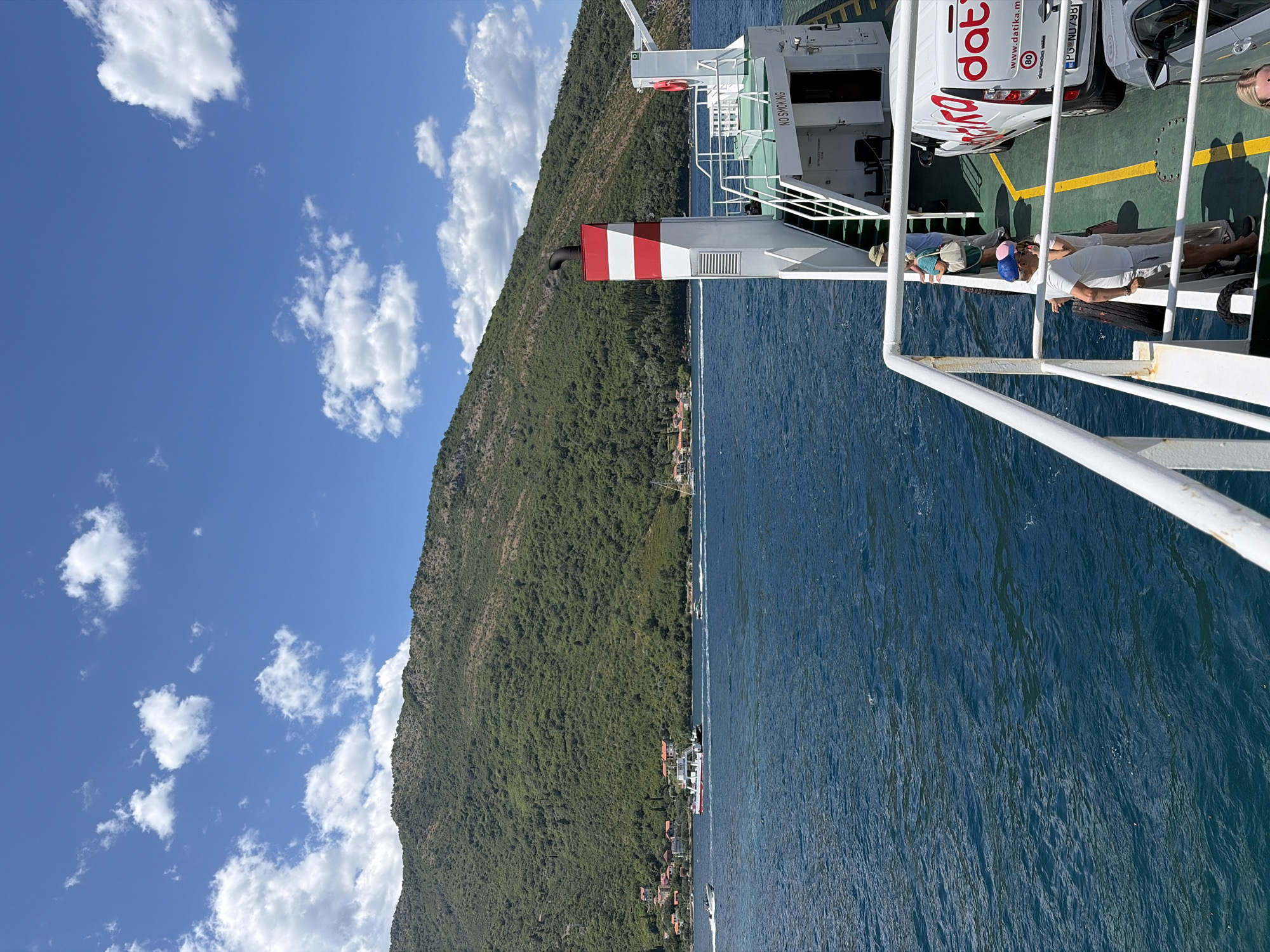 A ferry is sailing on a body of water with a lush green mountain in the background. The sky is bright blue with scattered white clouds. The ferry has a white structure with red and white stripes on top. Several people are visible on the deck of the ferry, some standing and looking out at the scenery, while others are seated. A white van with red lettering and a car are parked on the ferry deck. The water is deep blue and appears calm. Small boats are visible in the distance, navigating the water. The mountain in the background is densely covered with vegetation, and some buildings with red roofs are scattered along its base, suggesting a coastal town. The scene conveys a sense of travel and leisure on a sunny day. The location is the Strait of Verige in Lepetane, Montenegro, a common ferry crossing point.