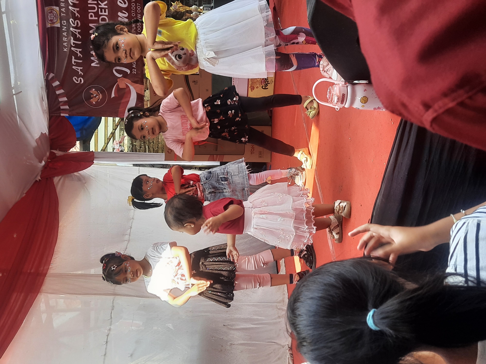 The image depicts a group of young girls performing on a stage in Bandung City, Indonesia. The stage is adorned with red and white drapery. The girls are dressed in colorful outfits, with some wearing sparkly makeup. They appear to be engaged in a dance or theatrical performance, with various poses and hand gestures. In the foreground, a person is seated, with their back to the camera, watching the performance. The background shows a banner with Indonesian text, likely related to the event. A water bottle is visible on the stage, near the performers. The overall scene suggests a community event or school performance taking place outdoors or in a semi-outdoor venue.