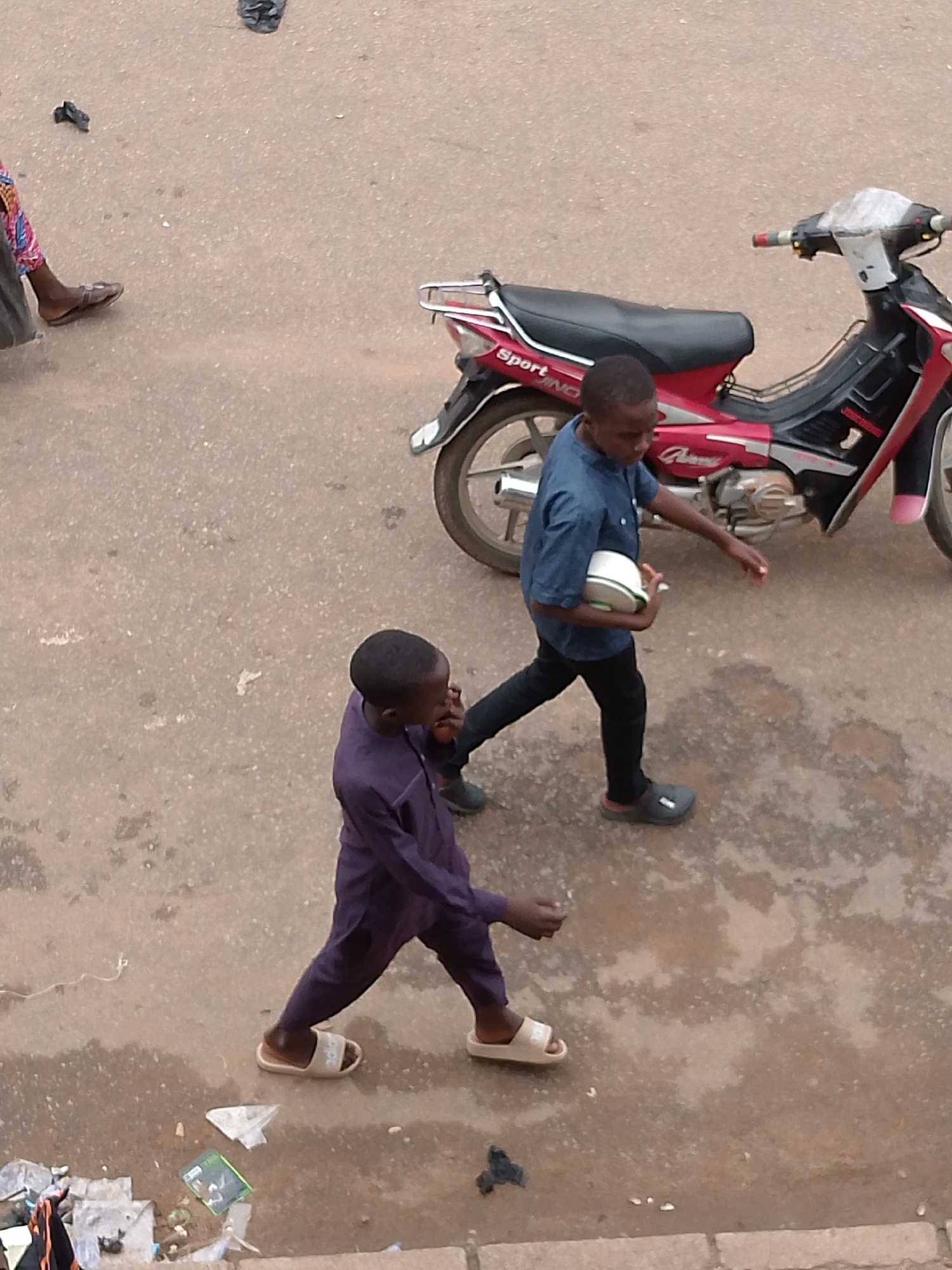 The image shows an overhead view of two young boys walking on a paved surface, possibly a street or sidewalk, in an urban environment. A red and black motorcycle is parked to the right of the frame. The boy on the left is wearing a dark purple robe-like garment and white slide sandals. He is walking towards the right side of the image, with his arms slightly outstretched. The boy on the right is wearing a blue short-sleeved shirt and black pants, with dark slide sandals. He is also walking towards the right, holding a white and green round object, possibly a ball or a container, in his left arm. He appears to be looking down at his hands. The ground is a mix of concrete and asphalt, with some wet patches and debris scattered around, including plastic bags and what appears to be discarded packaging. A person's legs, wearing a colorful patterned garment and brown sandals, are visible in the upper left corner, suggesting another pedestrian is present but mostly out of frame.