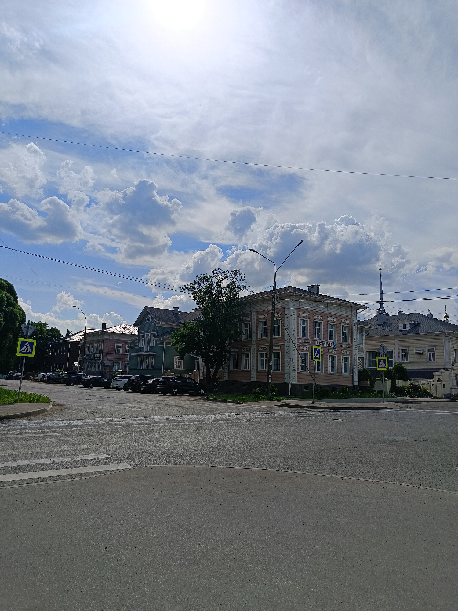 The image depicts a street intersection in Vologda, Russia, under a partly cloudy sky. In the foreground, there is an asphalt road with a pedestrian crossing marked by white stripes. To the left, a street sign for a pedestrian crossing is visible. Several parked cars line the street on the right side.
Behind the cars, a row of multi-story buildings is situated. The building closest to the right is a light-colored structure with a dark roof and a spire, possibly a church. Next to it is a three-story building painted in pink and white, featuring ornate architectural details. Further down the street, there are other buildings, one of which is painted green. The sky is predominantly blue with scattered white clouds, and the sun is bright, casting some glare.