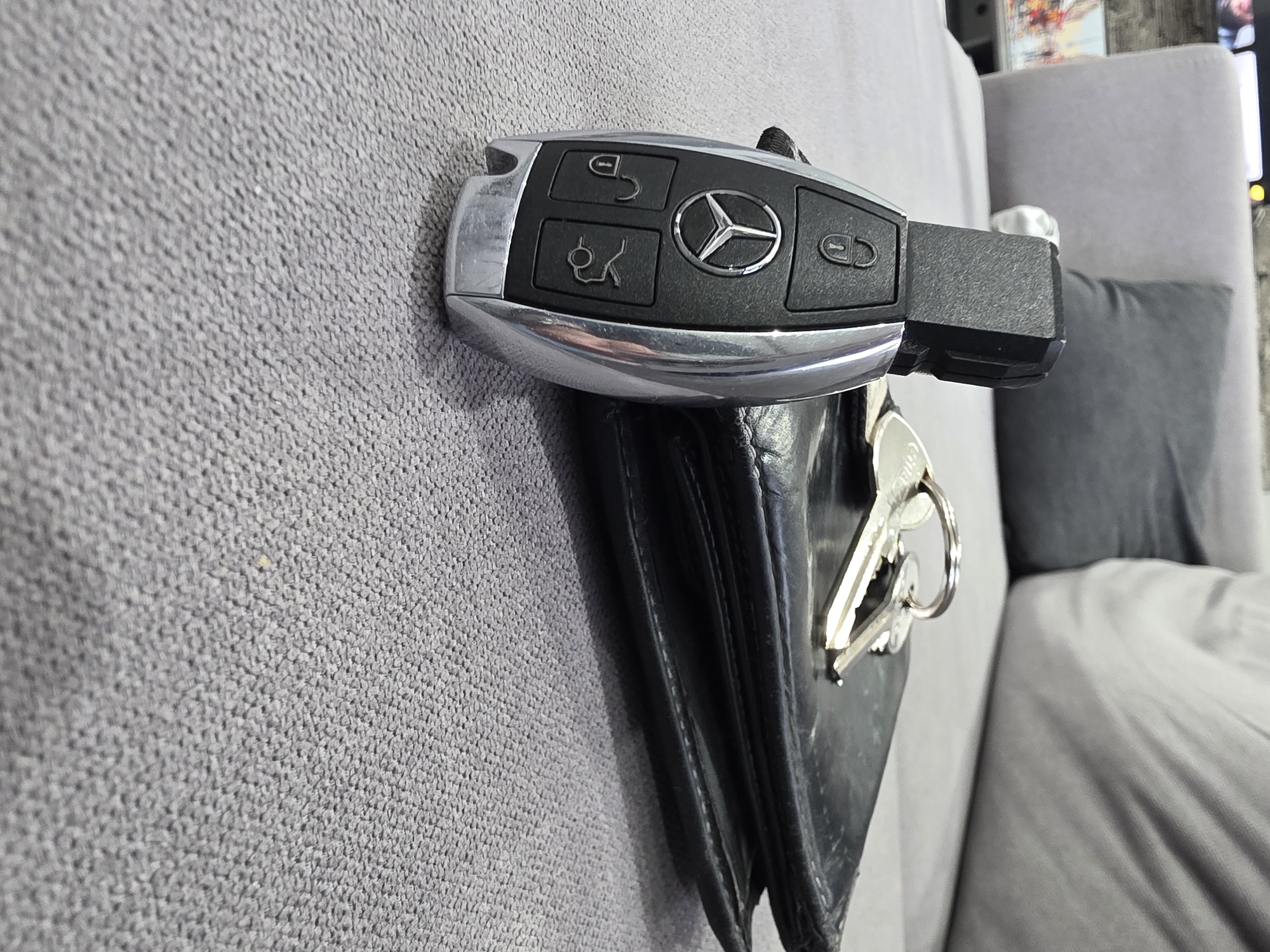 The image shows a Mercedes-Benz car key resting on top of a black leather wallet. Several other keys are also visible extending from the wallet. The objects are placed against a gray fabric surface, possibly a couch or other upholstered furniture. A gray pillow is partially visible in the background. The setting does not reveal information that would definitively place it in Yagodovo, Bulgaria. There are no people visible in the image.