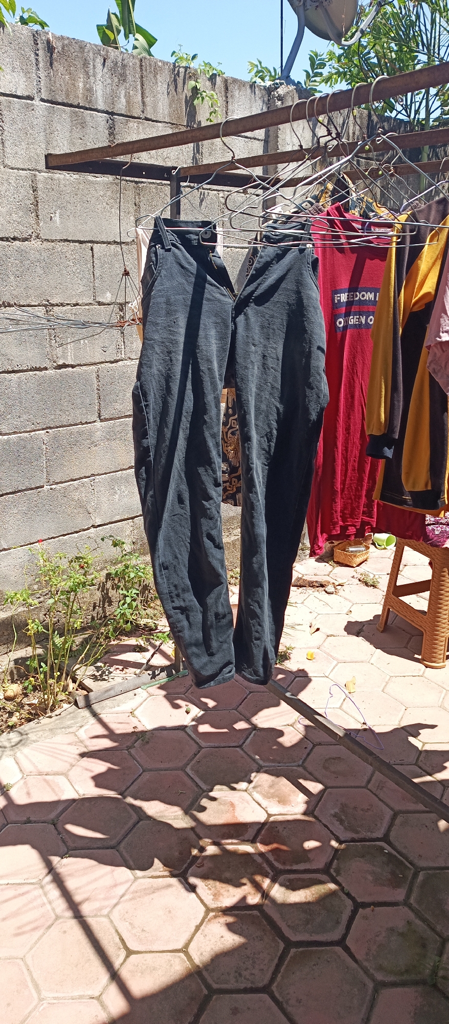 The image shows a close-up of a clothesline in an outdoor setting. Two pairs of dark-colored pants are hanging on the clothesline, along with several other garments that are partially visible. The clothesline is supported by a metal frame and is strung between two vertical posts, which are not fully visible in the frame. Behind the clothesline, there is a textured, gray concrete wall, possibly a building exterior. The ground is paved with hexagonal red tiles. Sunlight casts strong shadows from the clothes and the clothesline onto the tiled surface. Green foliage is visible at the bottom left and right edges of the frame. The sky appears clear and blue. The scene depicts a mundane, everyday activity of drying clothes outdoors.