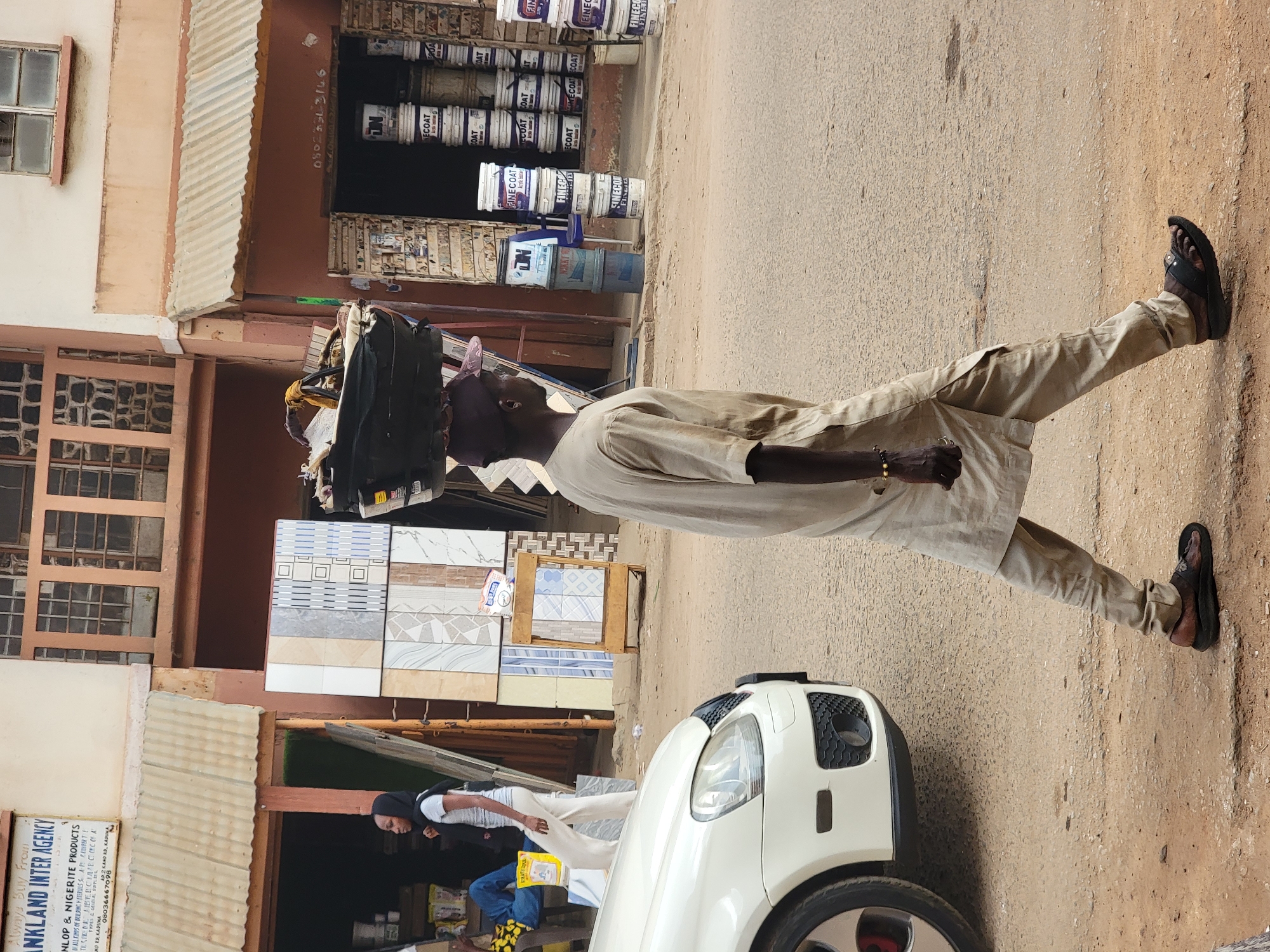 A man walks down a dirt road in what appears to be Maiburiji, Nigeria. He has items balanced on his head, possibly for transport or sale. The man is wearing light-colored traditional clothing and sandals. Behind him is a white car, and further back are buildings with various materials for sale, including paint buckets and tiles. Signs on the buildings advertise businesses like "ANKLAND INTER AGENCY" and "DUNLOP & NIGERITE PRODUCTS."