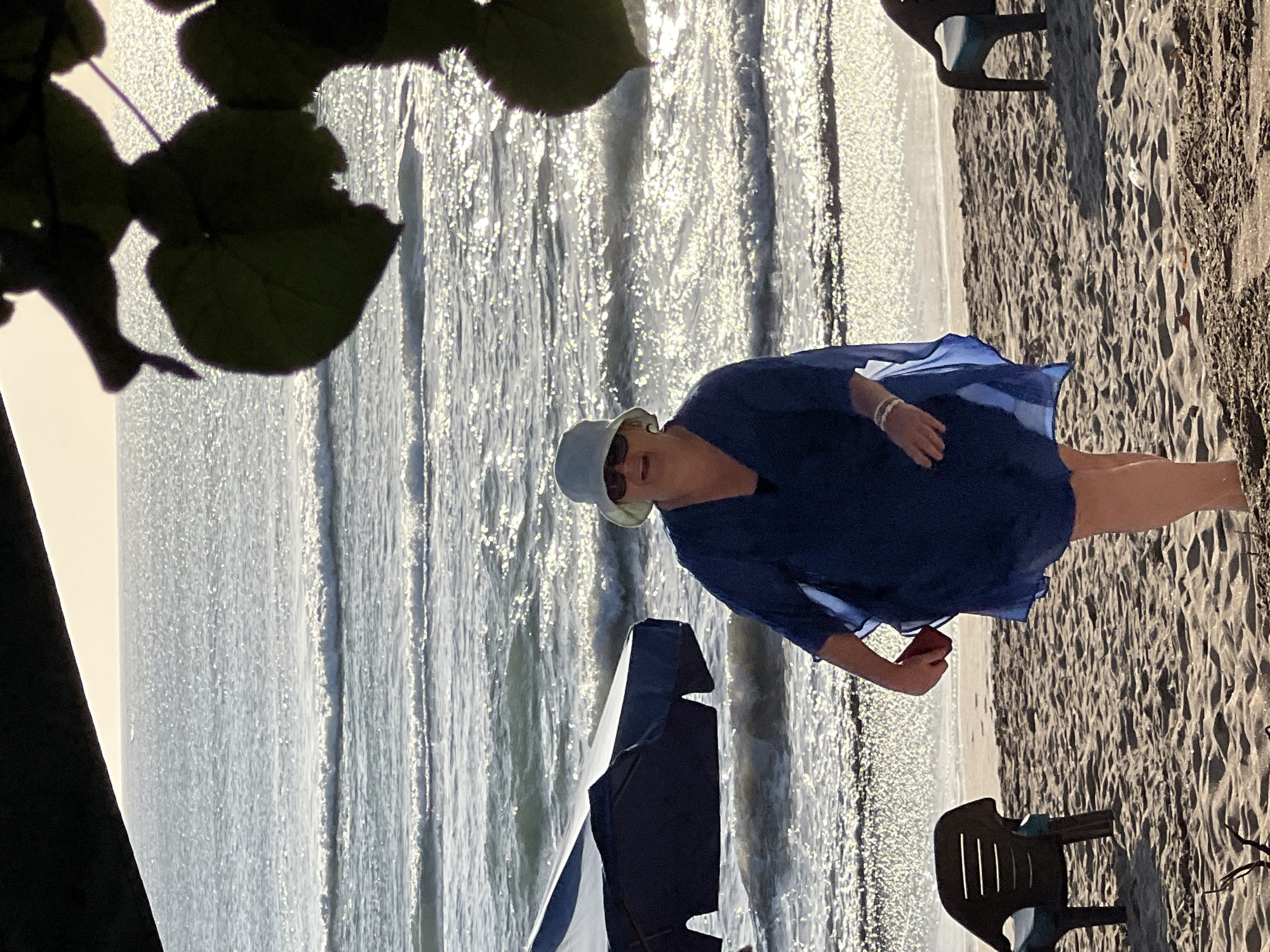 A medium shot captures a woman standing on a sandy beach, facing the ocean. She is wearing a blue cover-up, a light-colored hat, and sunglasses. Her right hand is holding a red object, possibly a phone. In the background, the ocean is visible with gentle waves and the sun is reflecting off the water. Two beach chairs and an umbrella are present on the sand. The scene is framed by foliage in the foreground, casting shadows. The lighting suggests it is either sunrise or sunset. The location is the beach in Kuta, Indonesia.
