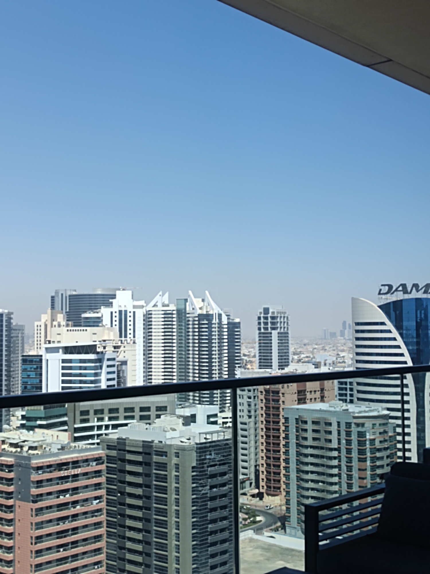 The image shows a cityscape in Dubai, United Arab Emirates, viewed from a high-rise building. The sky is clear and blue. In the foreground, there is a railing or balcony barrier. The mid-ground is dominated by numerous skyscrapers of various heights and architectural styles. Some buildings are white, others are clad in glass or patterned facades. A prominent building on the right has the word "DAMA" visible on its upper section. The background shows a hazy urban sprawl extending to the horizon, with more distant buildings visible. The lighting suggests it is daytime. There are no people visible in the image.