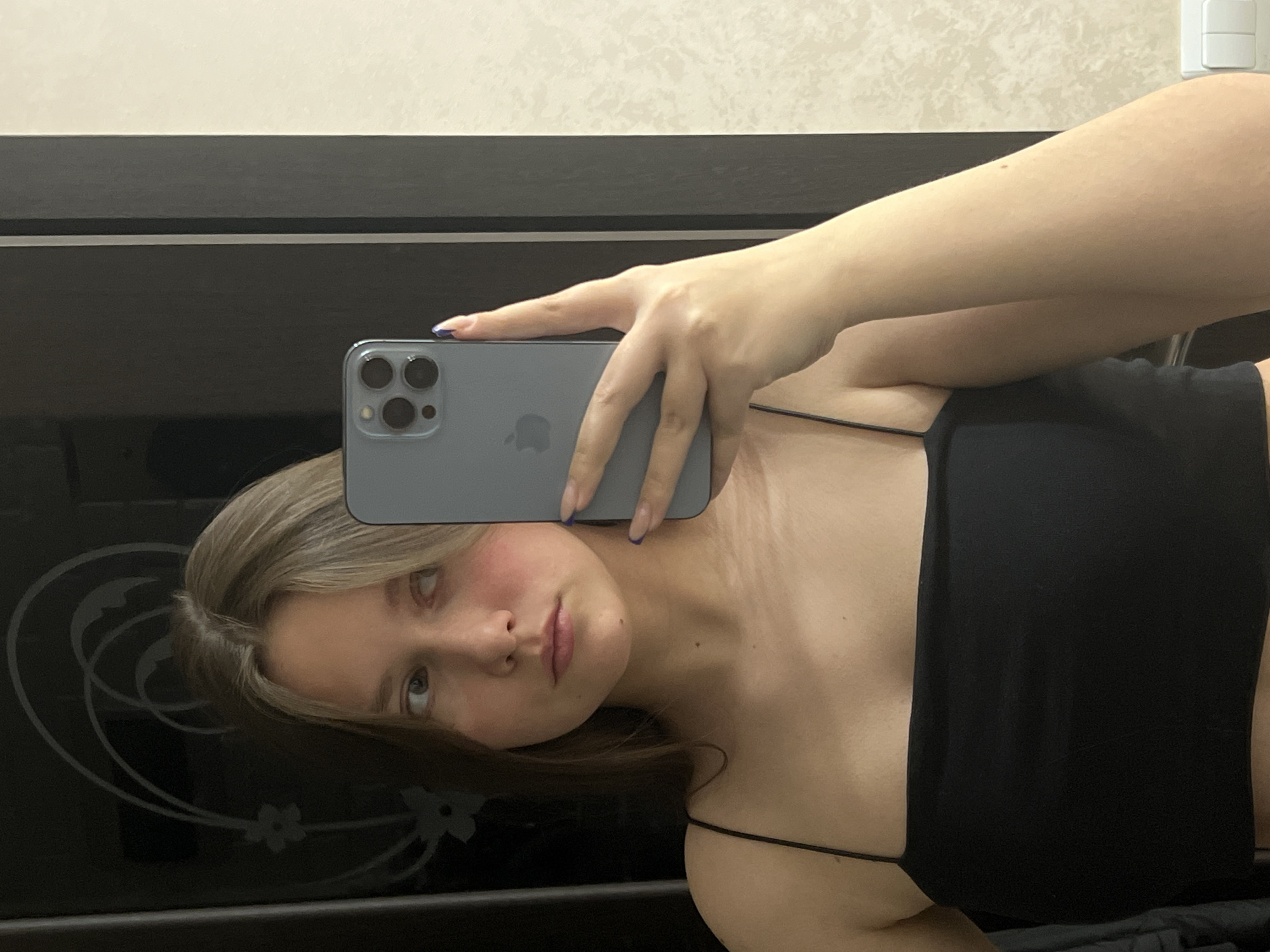 The image shows a young woman taking a mirror selfie indoors. The image is oriented with the woman's head to the left and her body extending downwards. She is holding a gray/blue iPhone, likely a Pro model due to its triple camera array and visible Apple logo, in her right hand. Her fingers, adorned with blue nail polish, grip the phone.
The woman has light brown hair and a neutral to slightly serious expression, looking towards her reflection. Her cheeks show a natural flush. She is wearing a black spaghetti-strap top.
The setting appears to be a room, possibly a bedroom, in a home. A dark, reflective surface, likely a piece of furniture such as a wardrobe or cabinet, occupies the lower part of the image and reflects part of her face and hair, including a faint floral or swirling decorative pattern. Above this furniture, a light-colored wall with a subtle textured pattern is visible. A white light switch is present on the wall in the upper right.
The woman is engaged in the act of photographing her reflection. There are no specific visual cues within the image to indicate the provided location context of Chaikovsky, Russia. The time of day is not discernible from the indoor lighting.