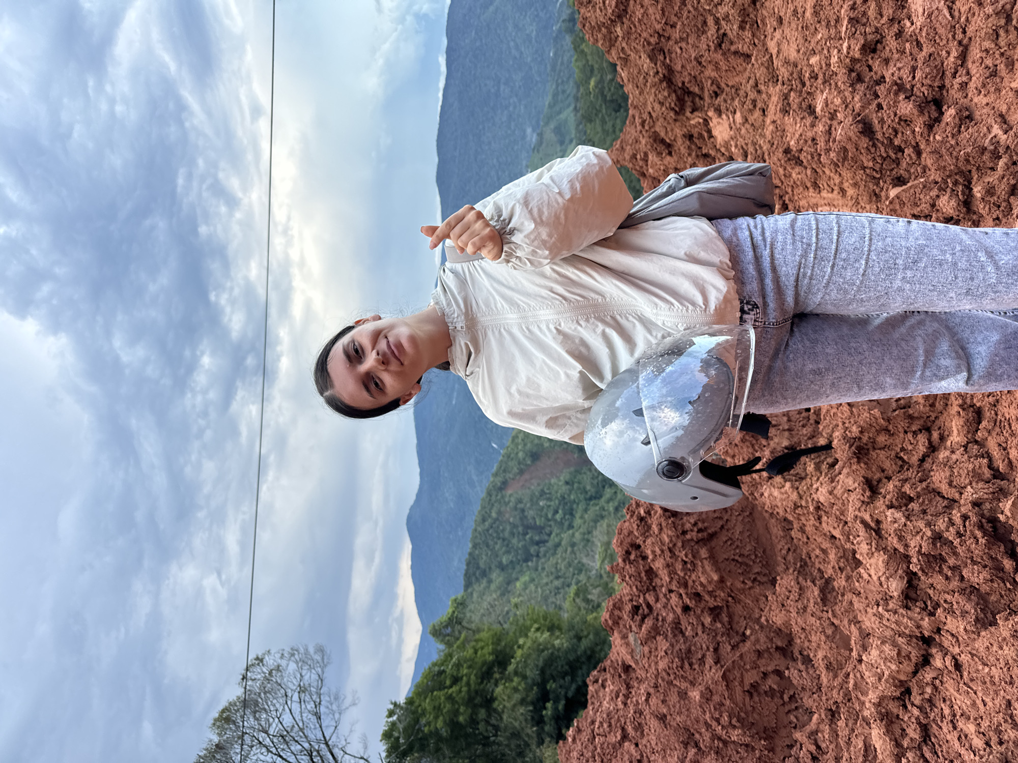 A young woman stands outdoors in a hilly, rural landscape, holding a gray motorcycle helmet in her left arm. She is wearing a light-colored zippered jacket and light-wash jeans, and has her right hand raised, forming a heart shape with her thumb and index finger. Behind her, the terrain is a steep, reddish-brown slope of loose soil, likely the result of a landslide. In the background, lush green mountains rise under a cloudy sky. The location is described as Nao Sẻ, Vietnam, suggesting a remote, possibly mountainous region.