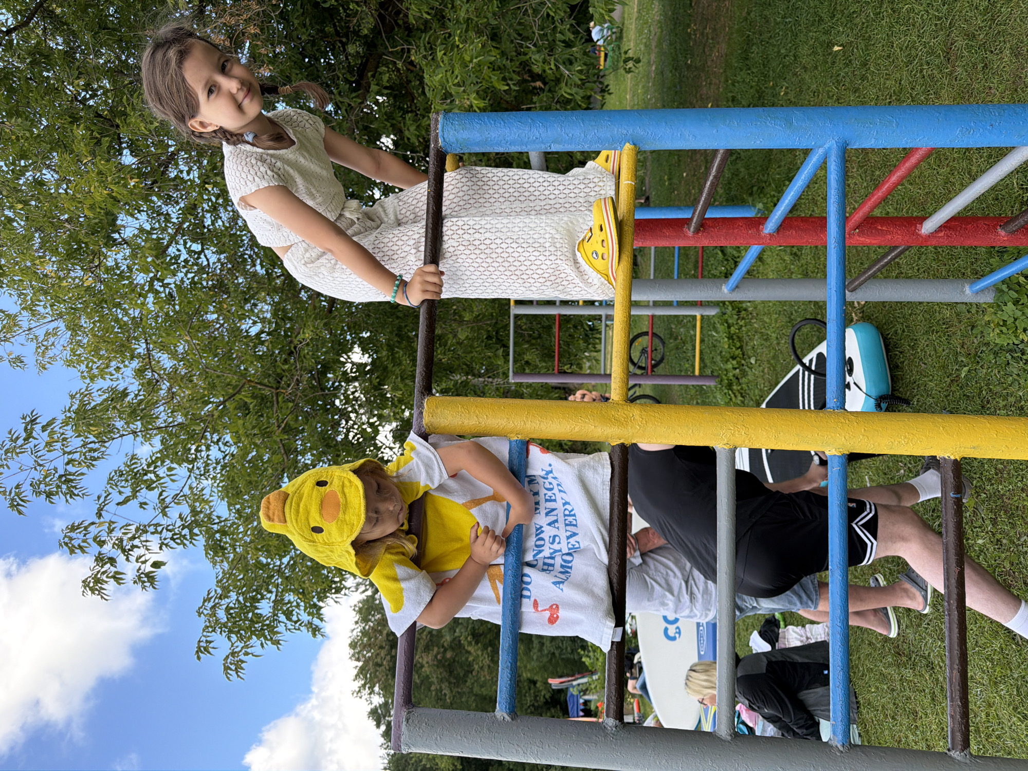 In Соколово, Russia, two young girls are playing on a colorful outdoor jungle gym on a grassy area with trees in the background. The girl on the left, wearing a yellow duck costume with a towel draped over her, is holding onto the bars, looking down. The girl on the right, in a white lace outfit, is leaning over the top of the jungle gym, looking towards the camera with a smile. Other people and objects, including surfboards and a bicycle, are visible in the background, suggesting a recreational event. The sky is blue with scattered clouds.