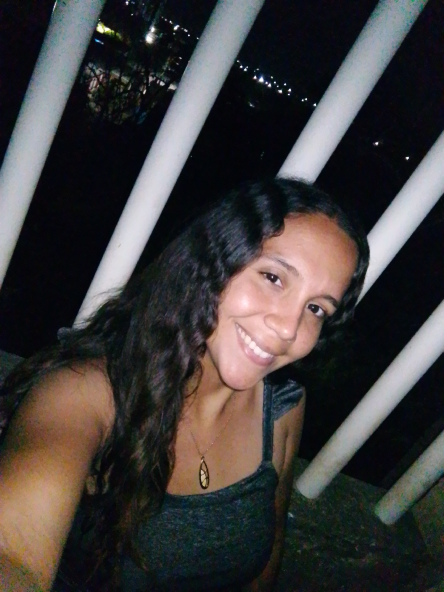 An image taken at night depicts an adult female subject positioned outdoors in Juan Griego, Venezuela. The subject occupies the lower-left to central portion of the frame, directly facing the camera with a broad smile. She has dark, wavy hair extending over her shoulders and wears a dark, sleeveless garment with thin straps. A gold-toned necklace with a tear-drop shaped pendant is visible on her chest. Her right shoulder and upper arm are prominent on the left side of the frame. In the background, several white, cylindrical bars, likely part of a railing or barrier, are angled diagonally across the scene. Beyond these bars, a dark environment reveals numerous small, bright lights, consistent with a city illuminated at night, viewed from an elevated perspective. Scattered dark shapes, possibly foliage or structures, are faintly discernible against the distant lights. The subject is directly illuminated, contrasting with the dark background.