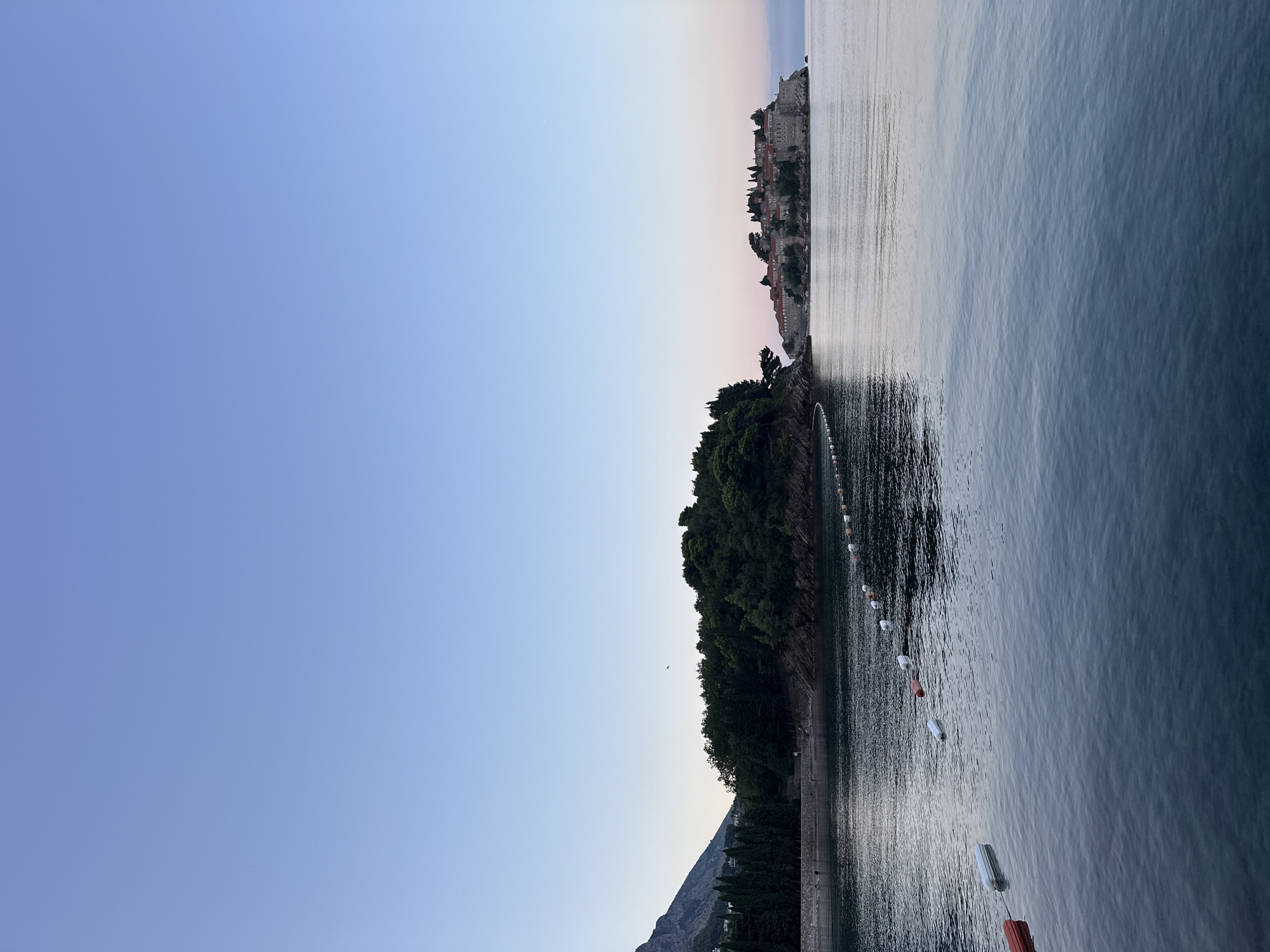 A wide shot shows the island of Sveti Stefan, Montenegro, with a calm sea in the foreground. The sky is a pale blue, transitioning to a soft pink near the horizon, indicating either dawn or dusk. The island, connected to the mainland by a narrow causeway, is covered with red-roofed buildings and lush green trees. In the water, a line of white and orange buoys marks a boundary. To the left, a forested hillside rises steeply from the water's edge, with the silhouette of a mountain visible in the distance. The water is a deep blue, reflecting the sky and surrounding land. No people are visible in the image.