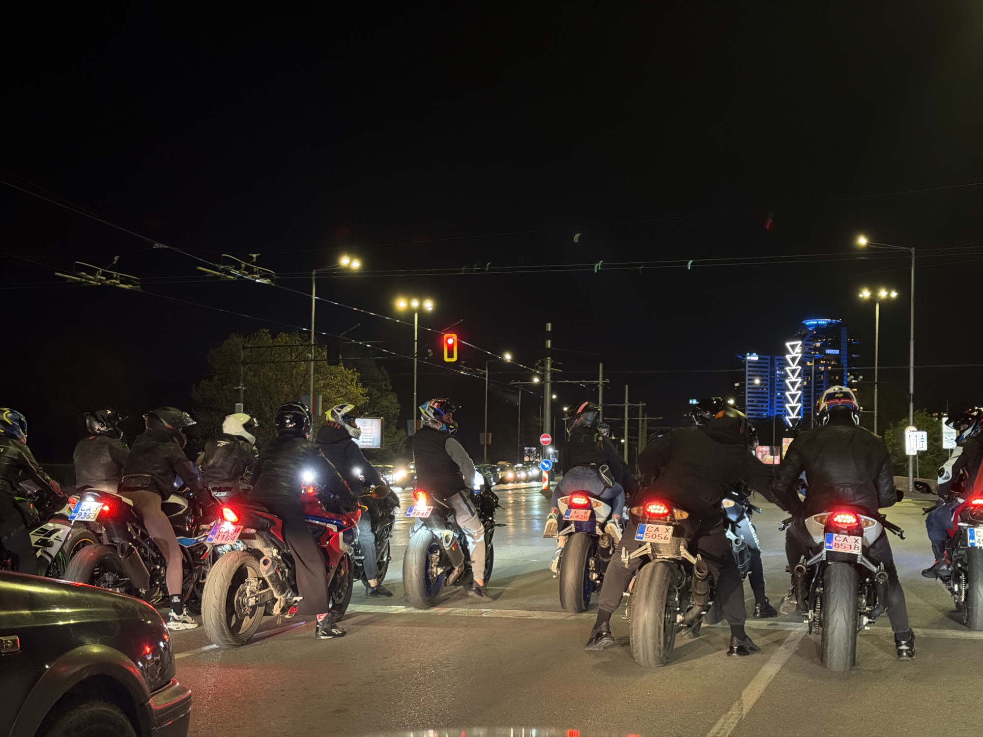 At night in Sofia, Bulgaria, a group of motorcyclists are stopped at a traffic light. Most riders are wearing helmets and dark clothing. The motorcycles are various models, with license plates visible on most. A partial view of a dark-colored car is in the lower left. Streetlights illuminate the scene, and a traffic light is visible ahead, showing red. A tall building with illuminated architectural details is in the background. Overhead wires and tram infrastructure are visible.