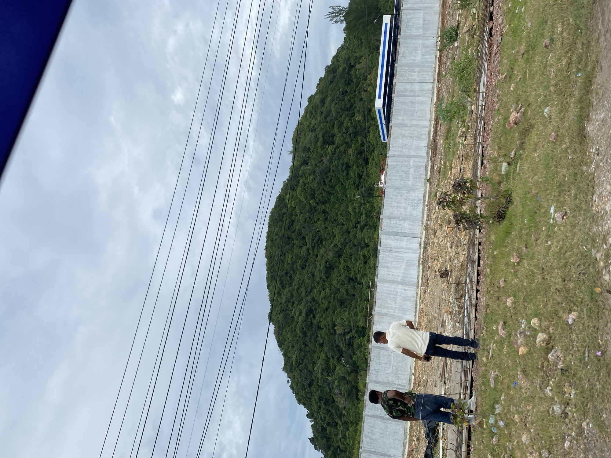 The image, taken near Lamkruet, Indonesia, shows two men standing near a construction site. A green, densely forested hill rises in the background. Overhead power lines are visible on the left side of the frame against a cloudy sky. A long, blue and white structure sits atop a concrete wall near the hill. The construction site is adjacent to a grassy area littered with rocks and debris. One man is wearing a white shirt and blue jeans, and the other wears a camouflage shirt and blue jeans.
