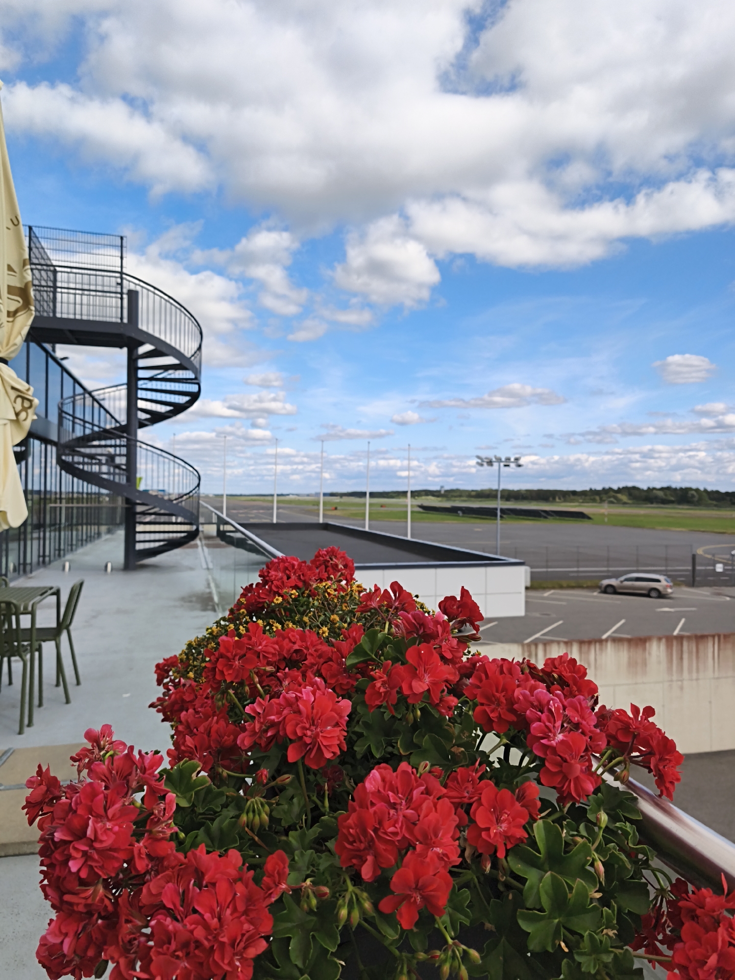 A medium shot captured from a slightly elevated vantage point shows a close-up of a lush planter filled with vibrant red geraniums in the foreground. Behind the flowers, a metallic railing is visible. Beyond the railing, a modern building with extensive glass windows and a dark, spiral staircase is positioned to the left of the frame. The building overlooks a paved area, likely a balcony or terrace, with a couple of dark green chairs and a table. Further in the background, an open expanse of an airport tarmac and runway stretches towards a tree line under a bright blue sky dotted with fluffy white clouds. A silver SUV is parked in a designated lot to the right of the building. The scene appears to be an outdoor seating area of a facility near an airport.