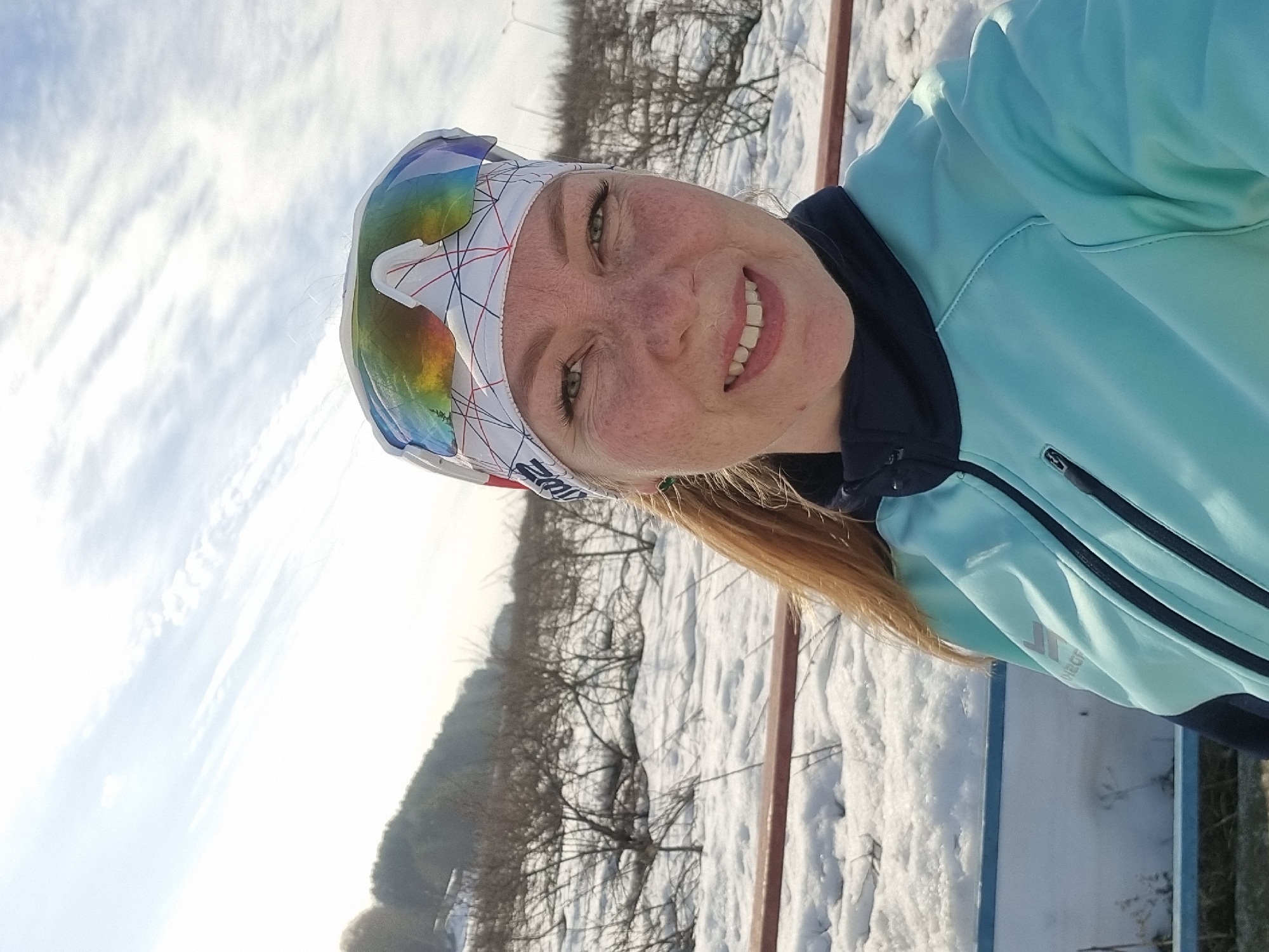 A selfie-style portrait captures an adult female outdoors in a snowy environment within Chaikovsky, Russia. The subject occupies the upper right portion of the frame, facing the camera directly with a smile that reveals her upper teeth. She has fair skin, visible freckles, and light-colored eyes. Her light brown hair is pulled back, with a ponytail visible on her left side. She wears a white athletic headband with red, blue, and black geometric patterns, topped with a pair of mirrored, multi-colored sports sunglasses. Her attire consists of a light blue/teal athletic jacket featuring dark blue trim on the collar and zipper lines. A partially visible logo is present on the left sleeve.
The background shows a snow-covered landscape under a bright, partly cloudy sky. Sparse, bare deciduous bushes and trees are visible in the mid-ground, with a forested, snow-dusted hill or mountain receding into the far background on the left. A reddish-brown horizontal railing or barrier runs across the middle ground behind the subject, beneath which a lower light blue/white structure is discernible. Distant light poles are faintly visible on the right horizon. The scene is illuminated by strong natural sunlight.