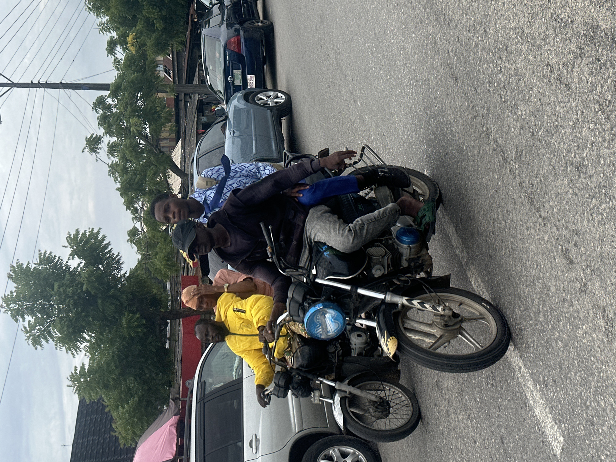 This image captures a busy street scene in Badagry, Nigeria. Several people are gathered around a motorcycle, with some riding on it and others standing nearby. The motorcycle appears to be overloaded, with at least three people on it, one of whom is driving. The people are dressed in casual clothing.
In the background, other vehicles are visible, including cars and what appears to be a bus, suggesting a lively urban environment. Trees line the street, adding a touch of greenery to the scene. The sky is overcast, indicating it might be a cloudy day. The overall atmosphere seems dynamic and perhaps a bit chaotic, typical of a bustling street in Nigeria.
One of the people on the motorcycle is wearing a yellow jacket, and another is wearing a blue and white patterned shirt with a blue tie. The motorcycle itself has a distinctive blue headlight cover. There is a pink umbrella visible on the left side of the frame. The ground is paved asphalt, with a white line marking part of the road.
The image appears to be taken during daylight hours, but the cloudy sky suggests it might not be midday with bright sunshine. The activity points towards daily transportation and possibly the movement of people within the town or to other destinations.