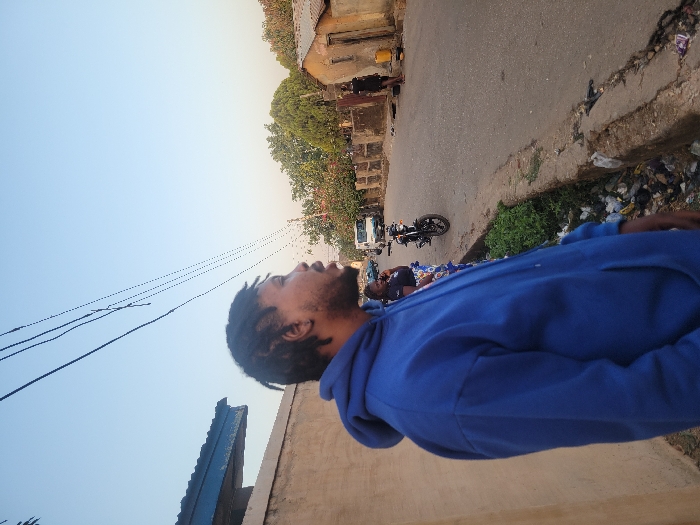 A man with dreadlocks, wearing a blue hoodie, gazes upwards. Overhead, power lines stretch across a bright, clear sky. Adjacent to the man is a building, and a street scene unfolds in the background. On the street, people gather near a motorcycle and a white van. The road borders a grassy verge and a wall with visible trash accumulation. Buildings line the street.