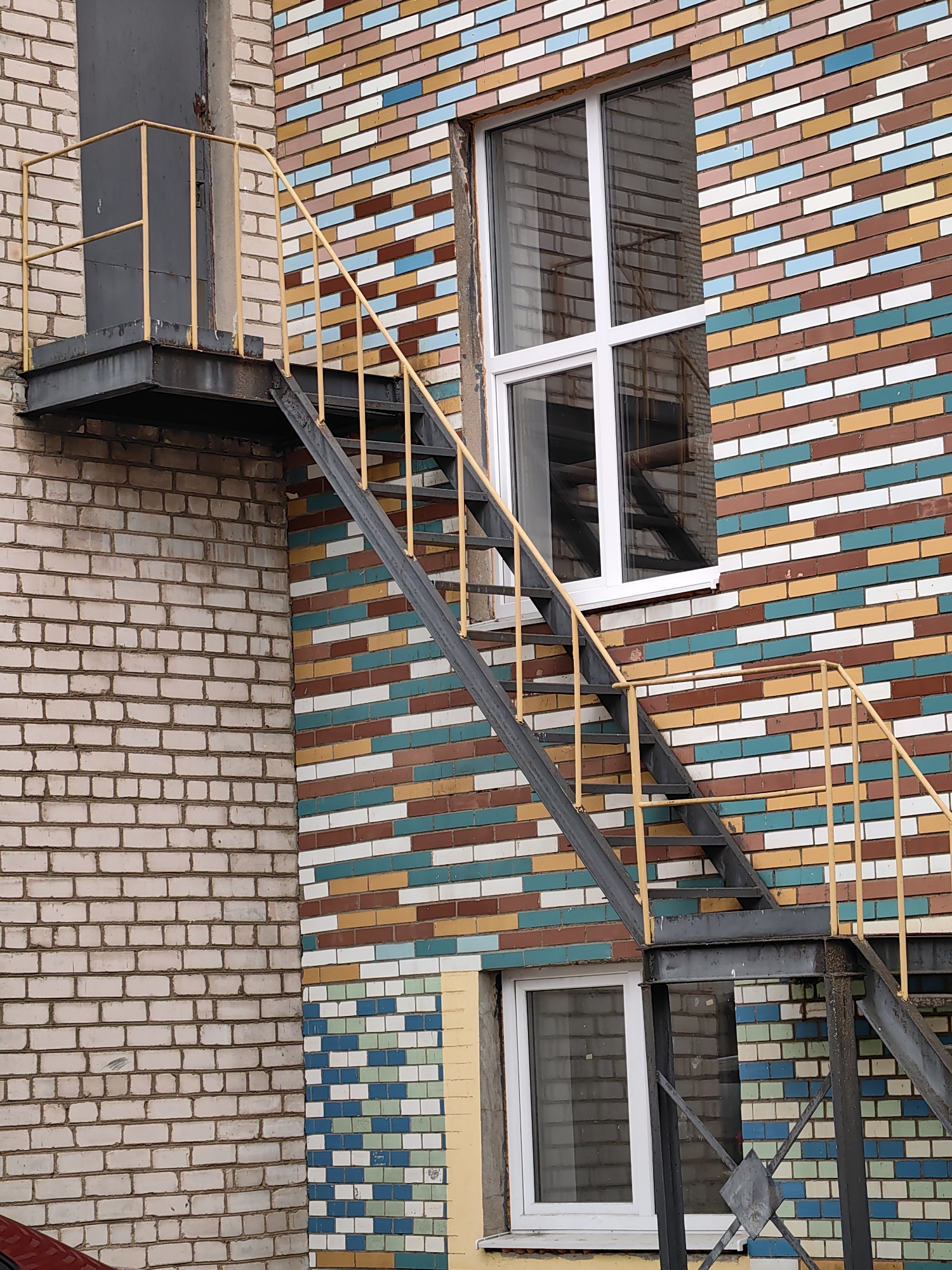 This image shows an exterior view of a building with a distinctive brick facade. The left side of the building is constructed with light-colored, roughly textured bricks. The right side features a wall covered in horizontal rectangular tiles of varying colors, including white, brown, tan, light blue, and teal, creating a colorful, patterned effect.
A dark grey metal staircase with yellow railings is attached to the tiled wall, leading upwards and to the left. The staircase appears to be an external fire escape or access route. A dark grey rectangular ventilation duct or pipe runs vertically along the left side of the building, near the staircase.
There are two white-framed windows visible. The upper window is larger and has a grid of four panes. The lower window is smaller and appears to be a single pane. Both windows have reflections of the interior, showing scaffolding or structural elements.
The lower part of the tiled wall features a different pattern of smaller square tiles in shades of blue, green, and white, arranged in a more geometric design. A section of a red vehicle is visible in the bottom left corner of the frame.
The lighting suggests it is daytime, possibly overcast, as there are no strong shadows. The setting appears to be an urban or industrial area, given the building's construction and the presence of external metalwork and a vehicle. There is no visible text in the image.