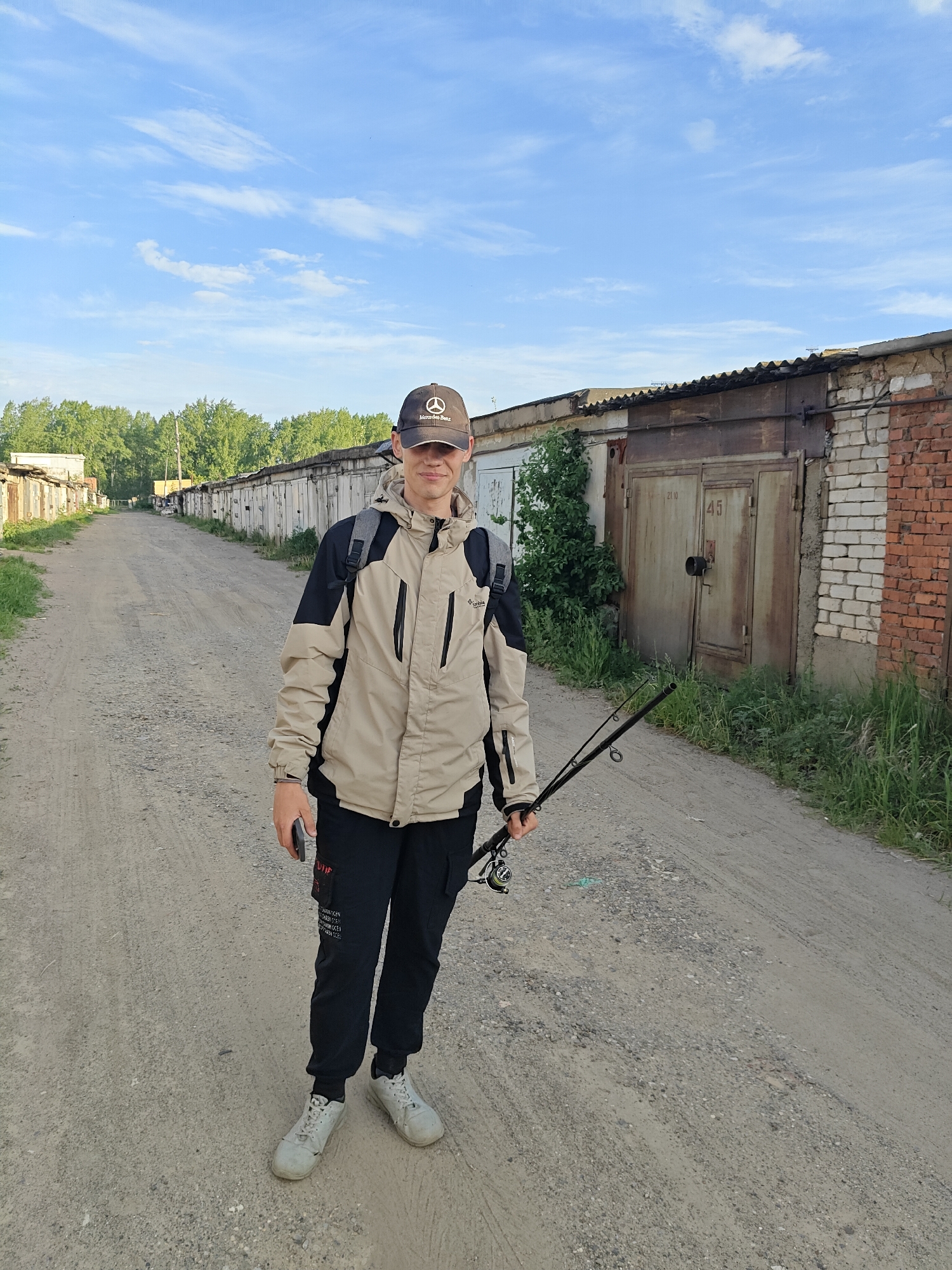 The photograph shows a young man standing in a dirt road in the city of Chaikovsky, Russia. He wears a Mercedes-Benz baseball cap, a tan and black jacket, black pants, and gray sneakers. He holds a fishing rod in his right hand and a cell phone in his left. He also wears a backpack. He is standing in front of a row of metal and brick garages, with vegetation growing nearby. The sky is blue with some clouds.