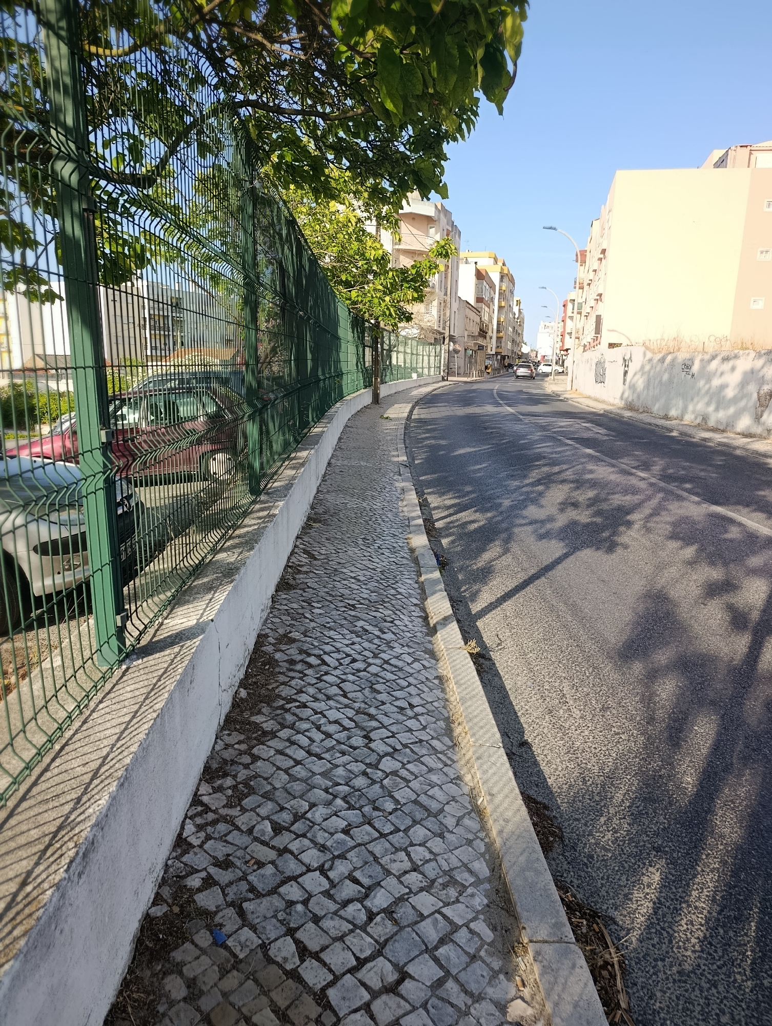 The image depicts a street scene in Setúbal, Portugal, with a sidewalk on the left and a street on the right. The sidewalk is paved with cobblestones and is bordered by a concrete wall and a green metal fence. Beyond the fence, several cars are parked, and trees with green leaves are visible. The street itself is asphalt and appears to be a two-way road with a white line marking the center. Buildings line the right side of the street, some of which are residential apartments and shops. The sky is clear and blue, indicating a sunny day. Shadows of trees are cast across the street and sidewalk, suggesting the sun is positioned to the right and slightly behind the viewer. There are no people or animals visible in the frame. The overall impression is one of a quiet, daytime urban environment.