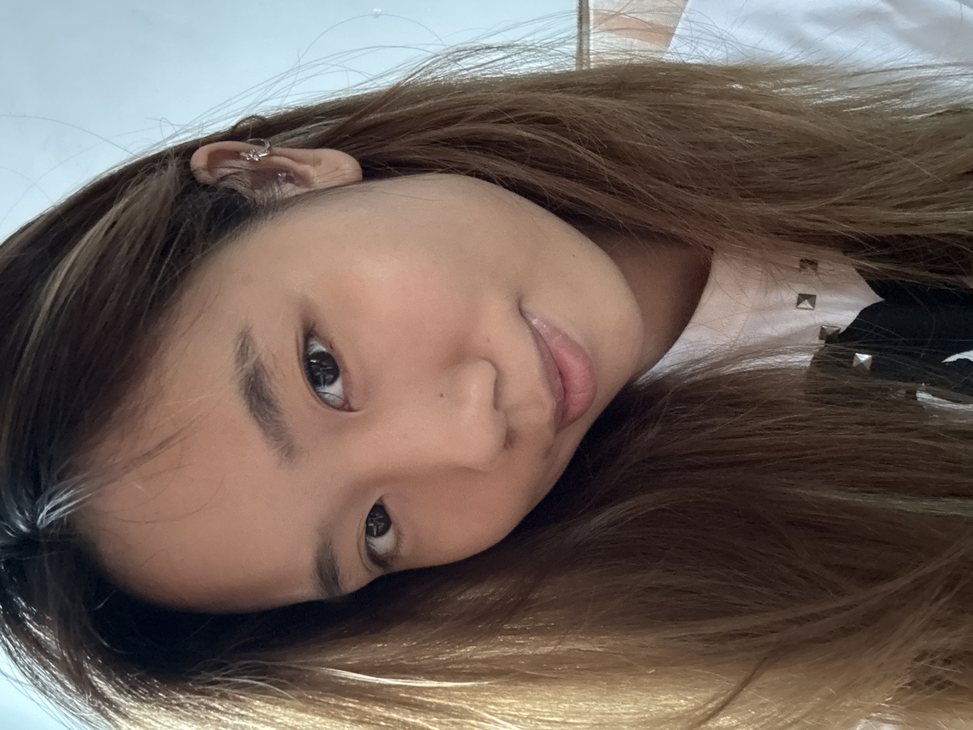A close-up, chest-up selfie of a young woman with medium-length, straight brown hair, some of which frames her face. She is looking towards the camera with a slight smile. She is wearing a white t-shirt with silver stud accents and a black design on the front. The woman has a small stud earring in her right ear. The background is out of focus and appears to be an indoor setting with a light blue wall and a hint of a striped pattern. The image quality is sharp, with good lighting. The image was taken at a high angle, likely by the subject herself, and the setting does not provide any specific indicators of its location within Guandu, China.