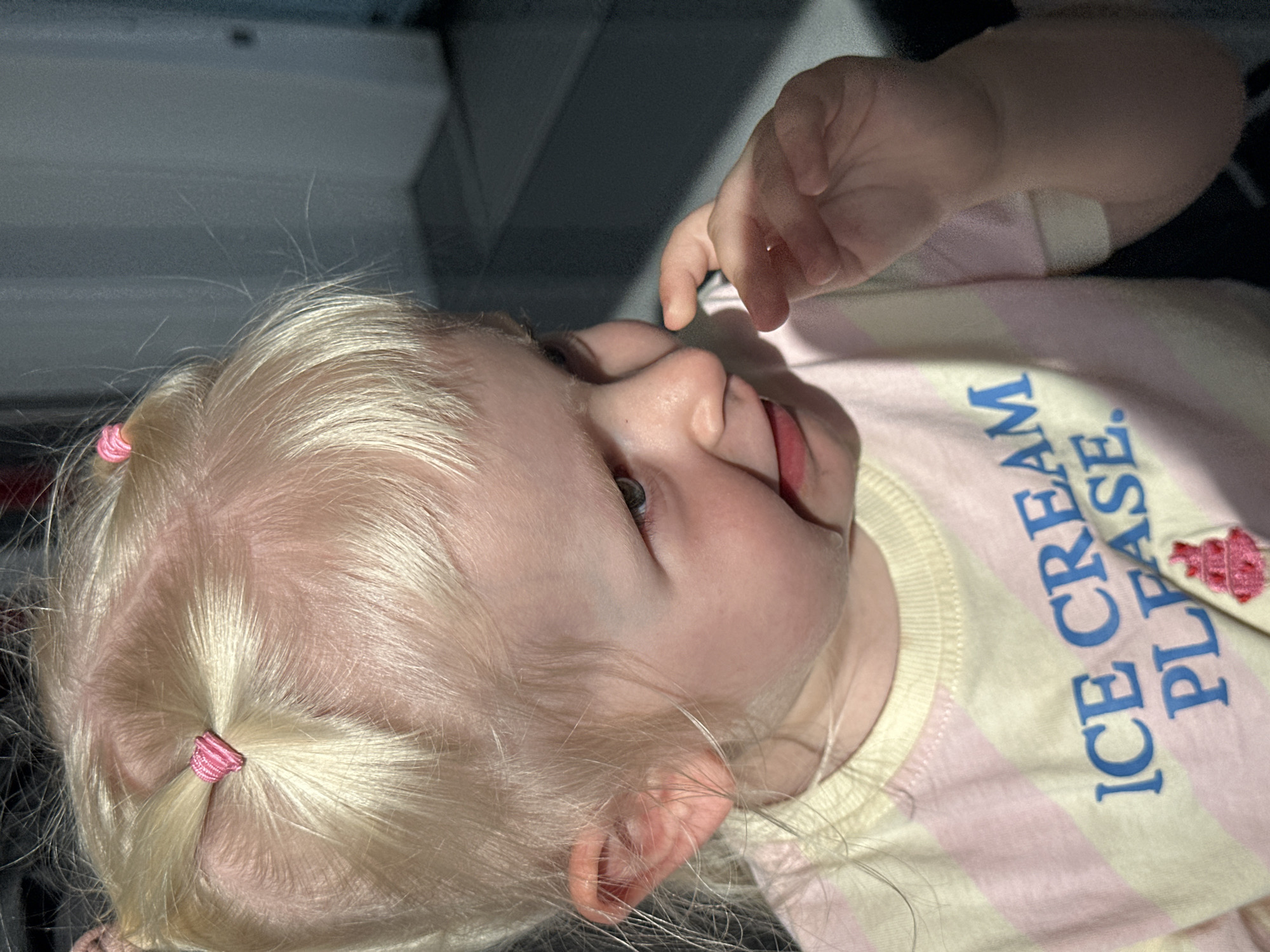 A fair-skinned, blonde-haired toddler is angled in the center of the frame. The child's hair is pulled into two small ponytails, secured with pink hair ties. The toddler's right hand is raised, fingers extended. The child wears a light-colored striped shirt with the words "ICE CREAM PLEASE." The background is blurred and features gray and black tones. This image does not provide enough information to verify that the location of the image is in the city of Petah Tikva, Israel.